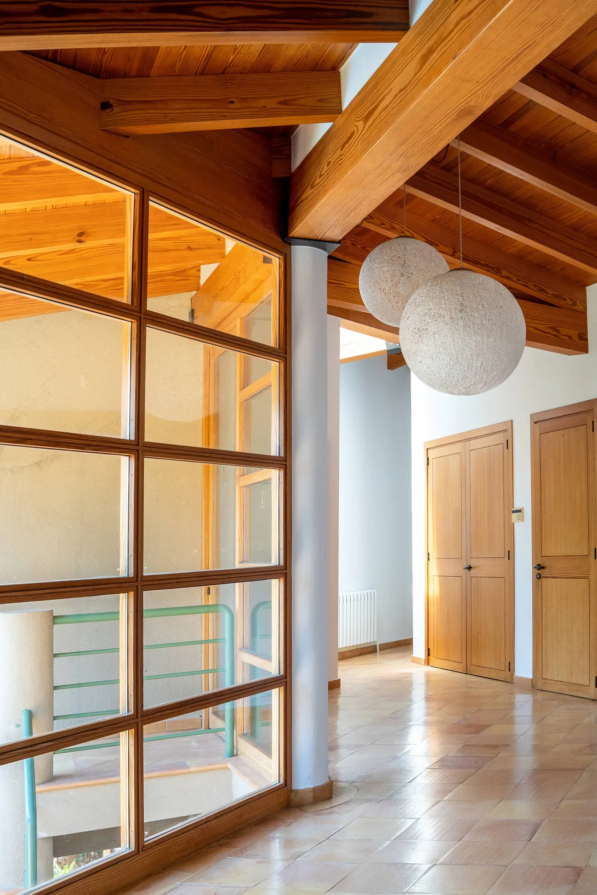 Un pasillo interior luminoso con vigas de madera, paredes acristaladas y puertas de madera.