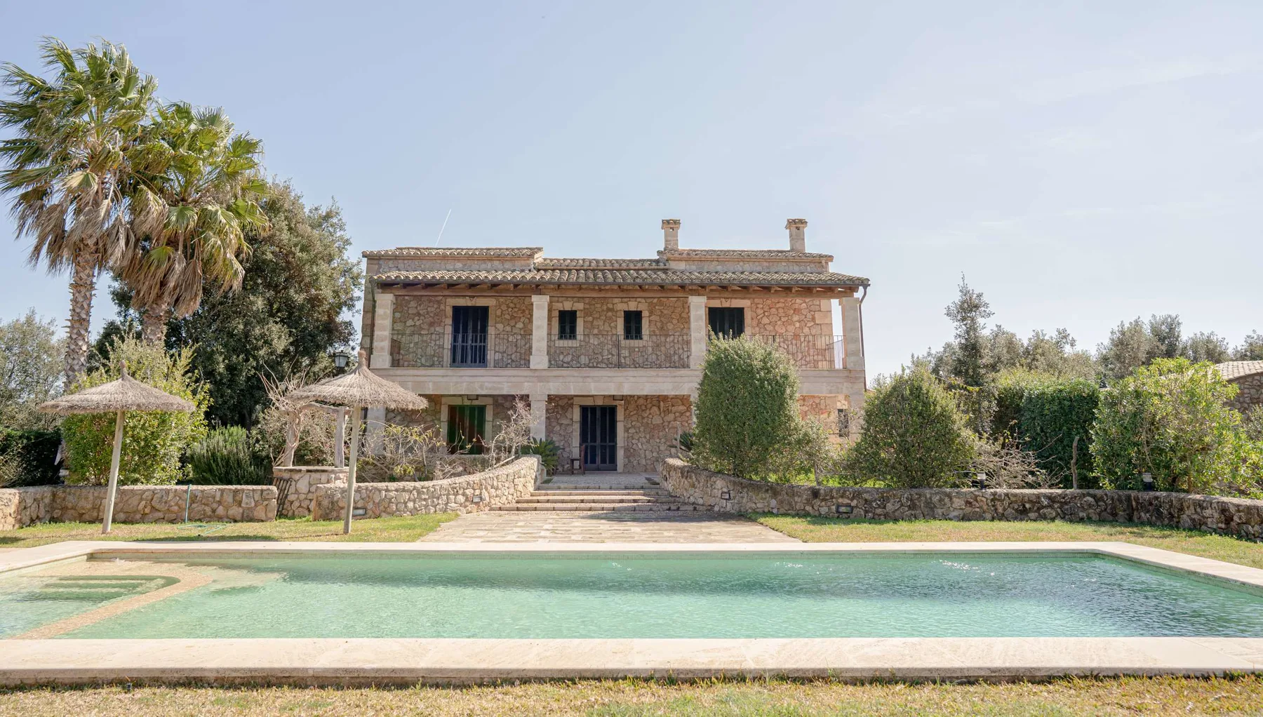 Una casa de piedra se alza detrás de una piscina, con árboles y sombrillas cerca.