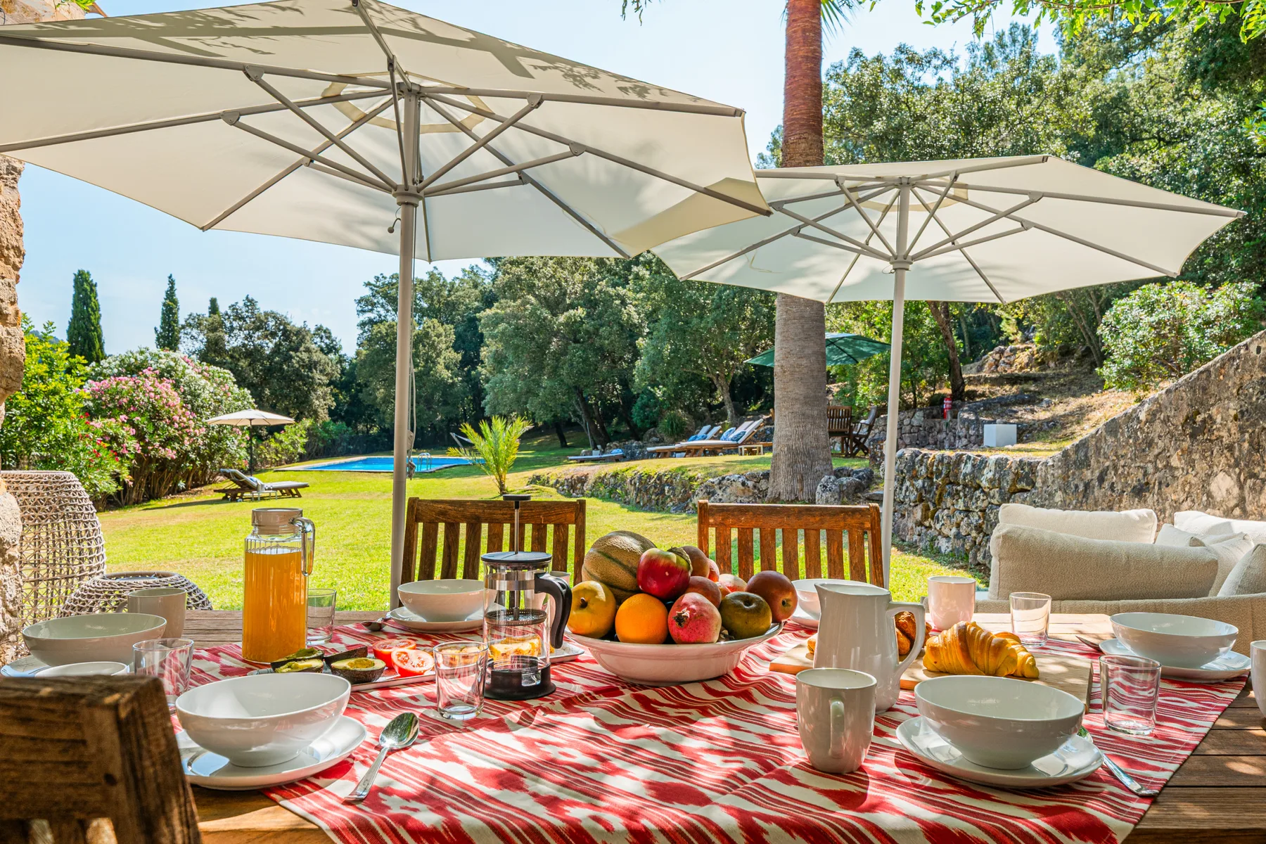 Una mesa de comedor al aire libre está preparada bajo sombrillas junto a un jardín.