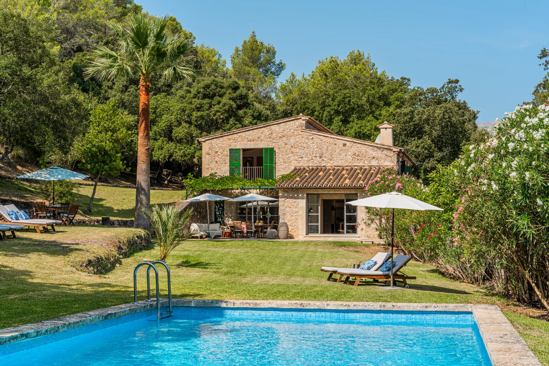 Una casa de piedra da a una piscina, con tumbonas sobre el césped.