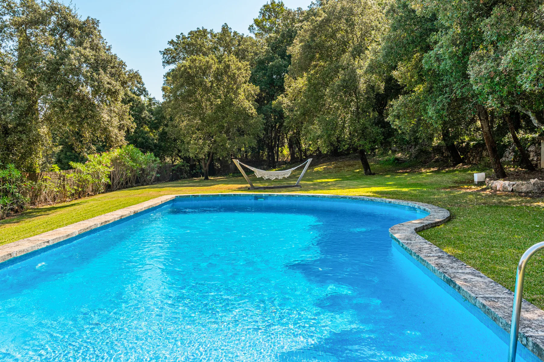 Una piscina al aire libre rodeada de árboles, con una hamaca en el césped.