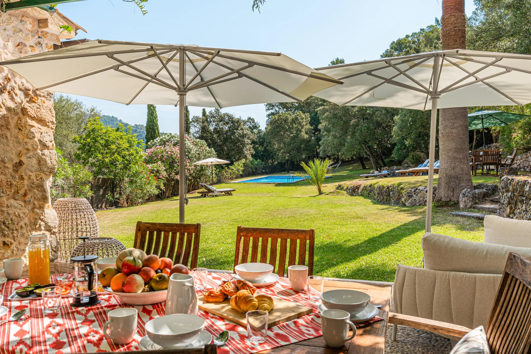 Comedor en el jardín con una mesa puesta bajo sombrillas, frente al césped y la piscina.