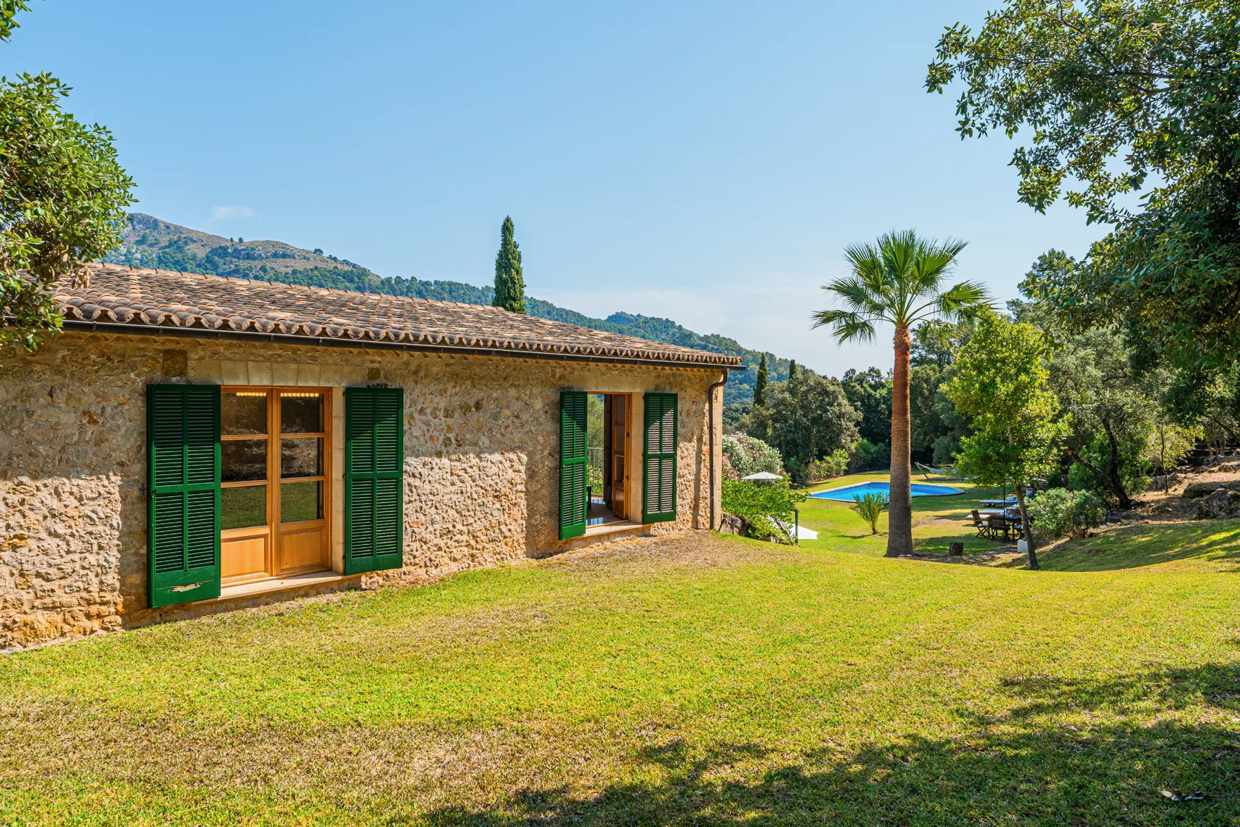 Una casa de piedra con contraventanas verdes está junto a un césped soleado y un jardín.