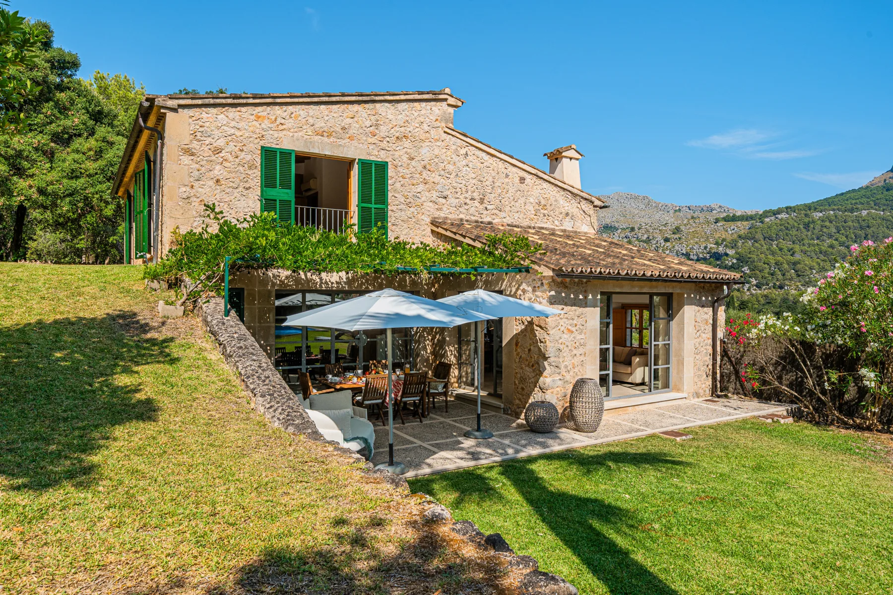 Una casa de piedra da a un patio con sombrillas y un jardín con césped.