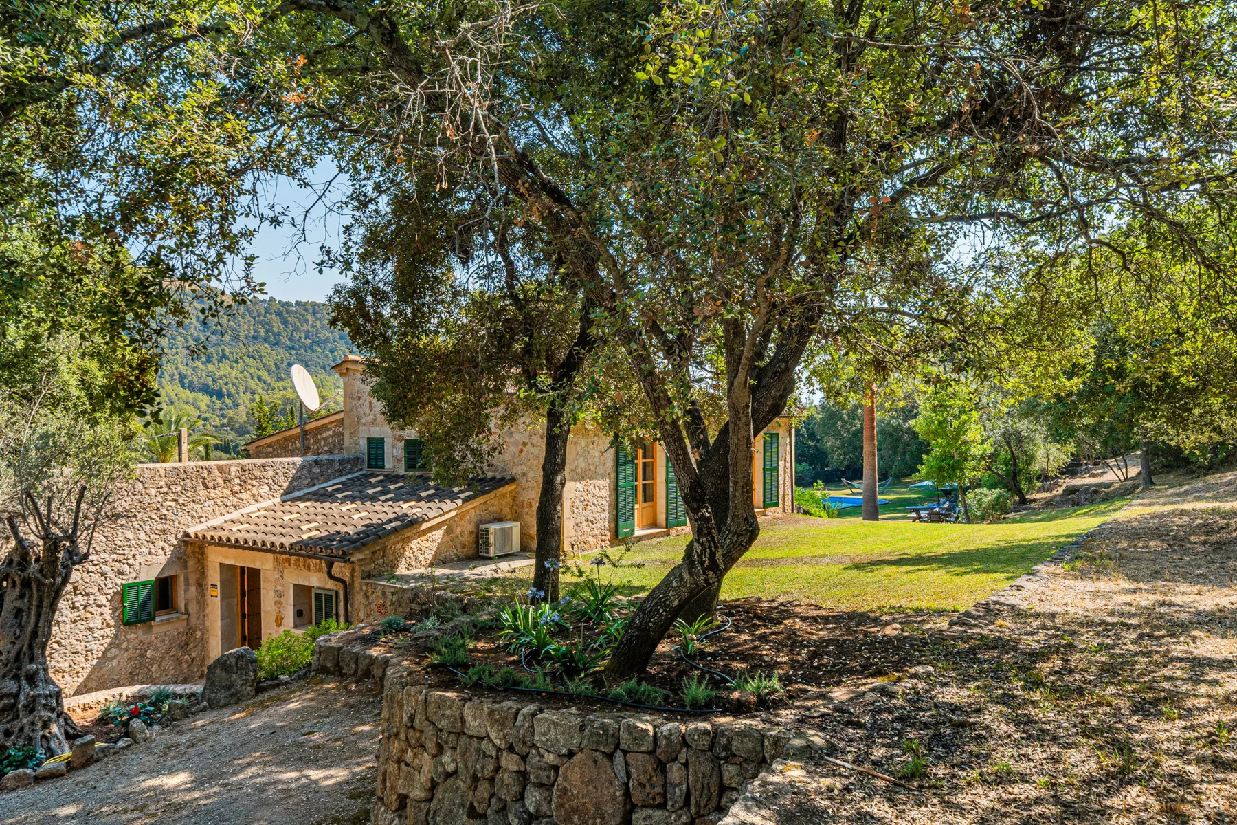 Una casa de piedra está junto a un jardín sombreado con árboles maduros.