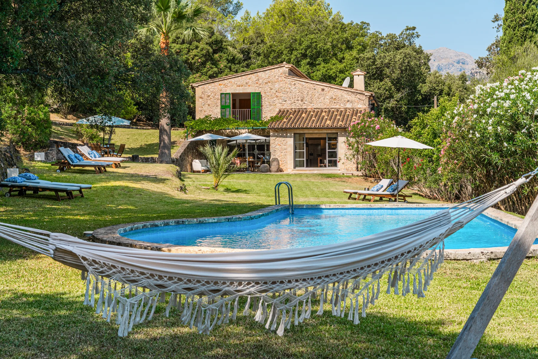 Una piscina y una hamaca están frente a una casa de piedra en un jardín verde.