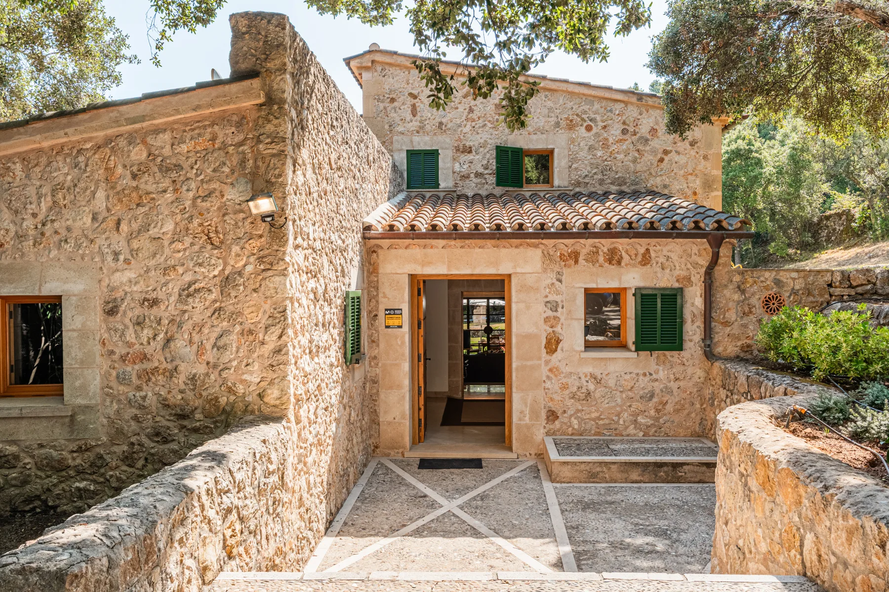 La entrada de una casa de piedra da a un patio pavimentado bajo árboles frondosos.