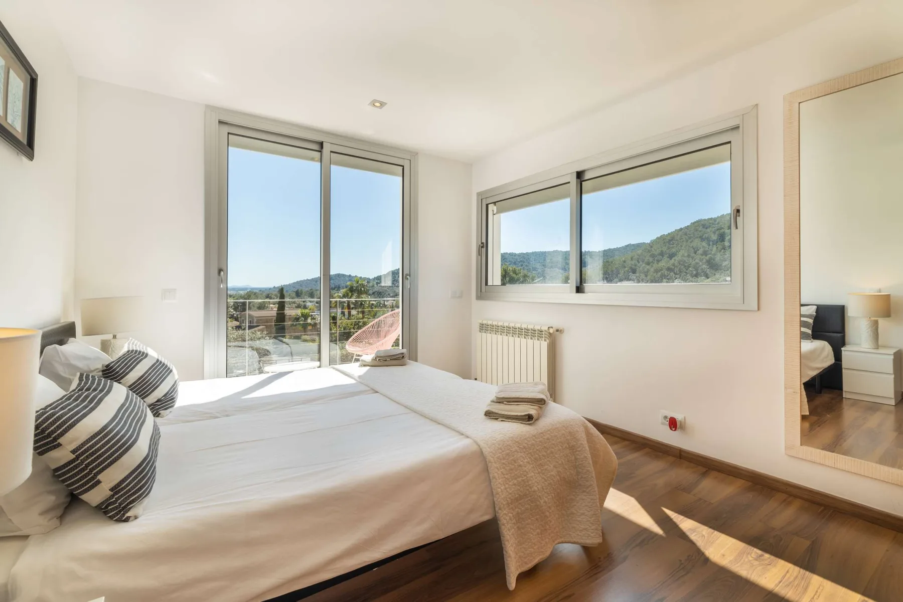 A bright bedroom with a made bed beside large windows overlooking green hills.