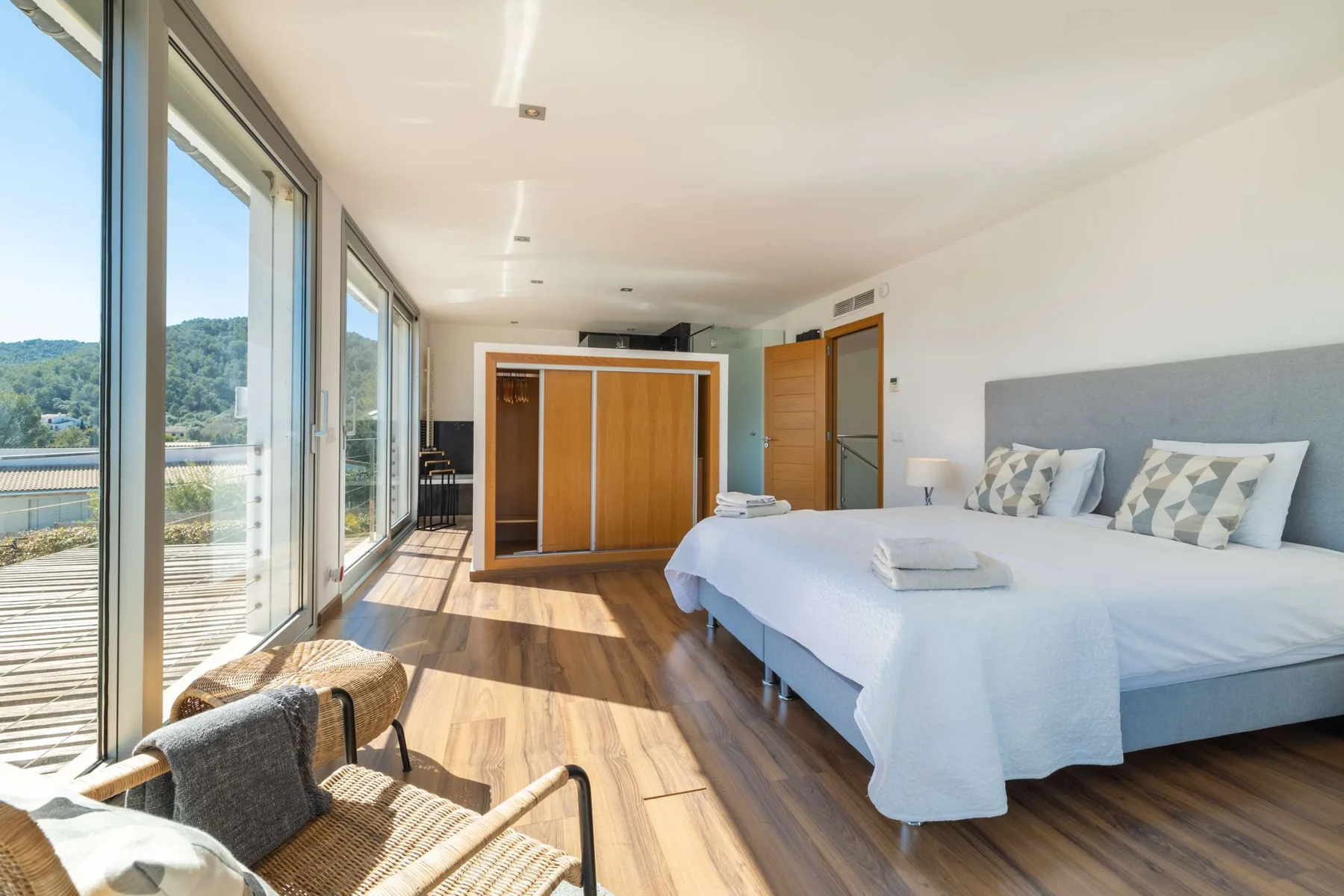 A bright bedroom with beds, a wardrobe, and large windows overlooking greenery.