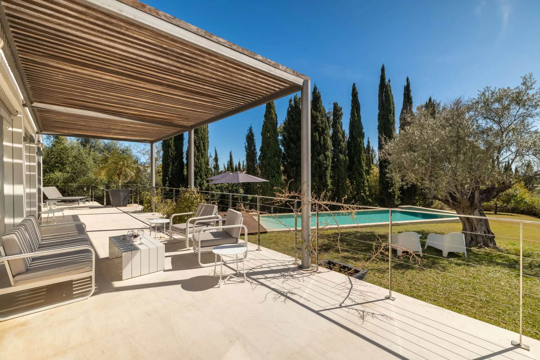 A poolside patio with shaded seating and a view of tall trees.