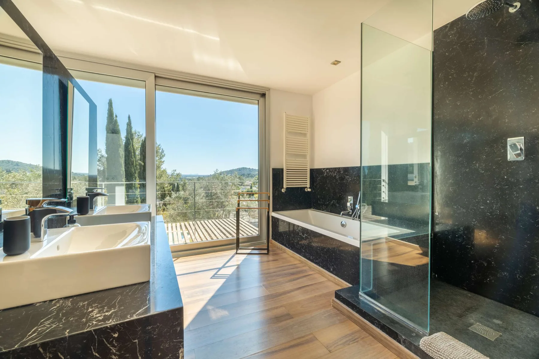 A bright bathroom with a bathtub, glass shower, and large windows overlooking greenery.