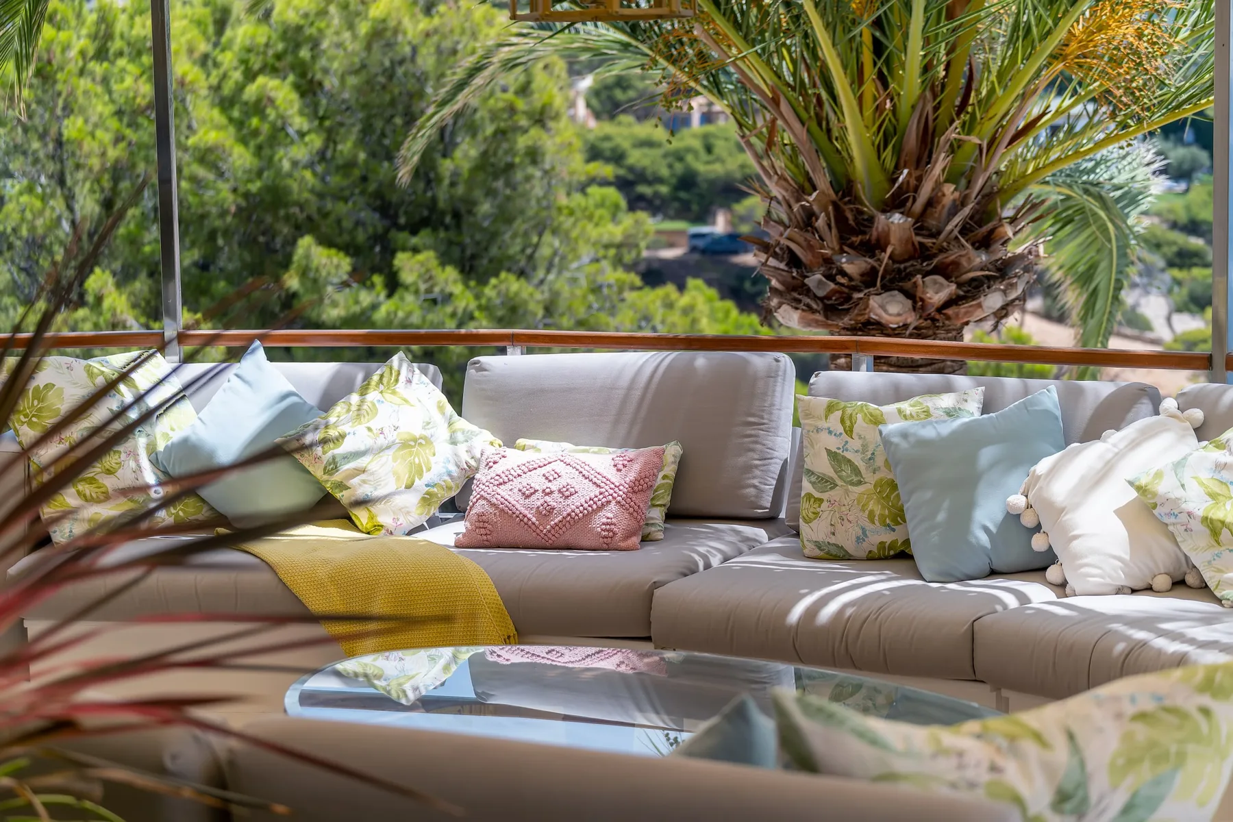 A cushioned outdoor seating area sits beside a glass railing, surrounded by greenery.