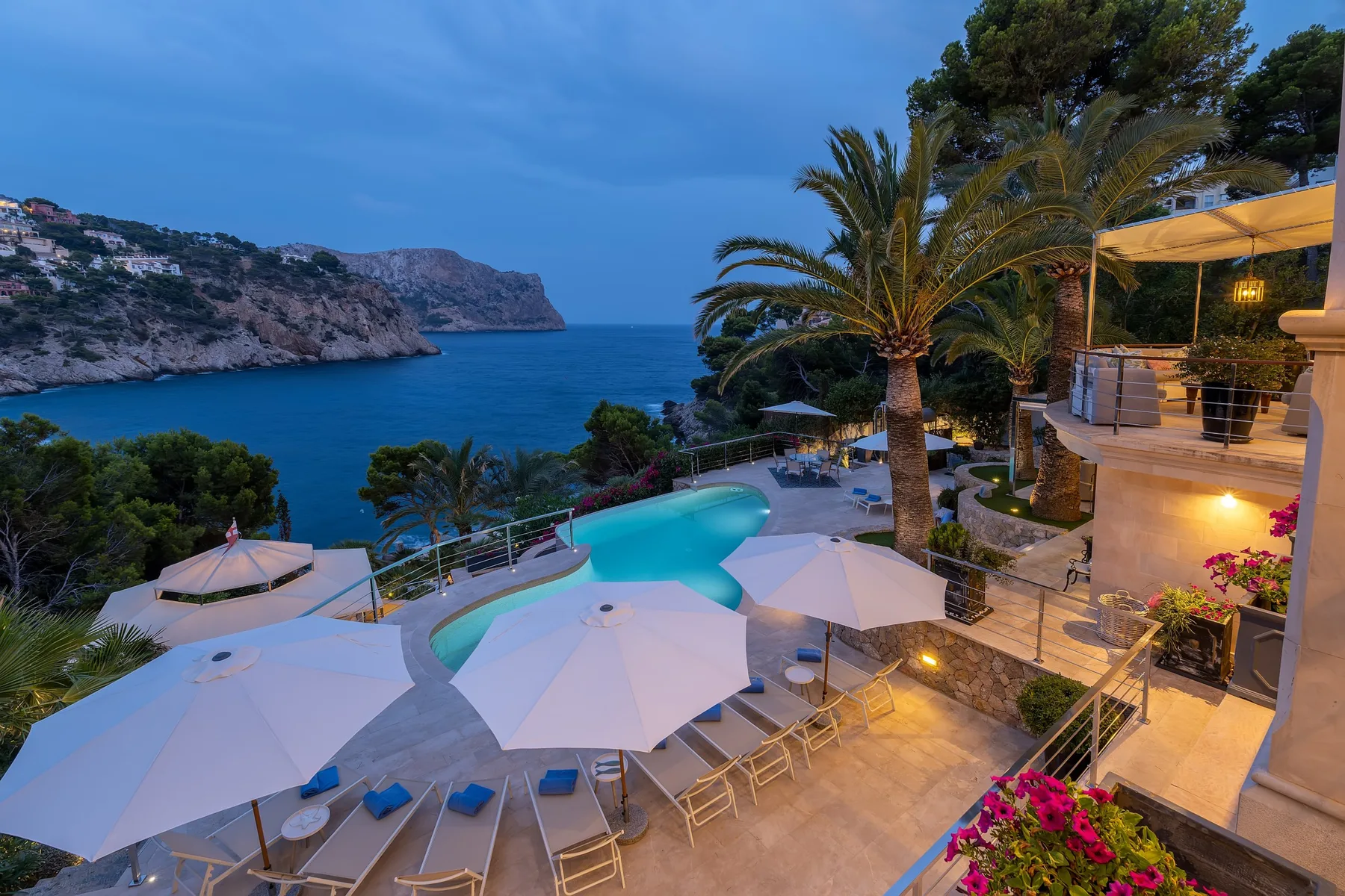 A seaside terrace overlooks a coastal bay, with a pool and shaded seating.