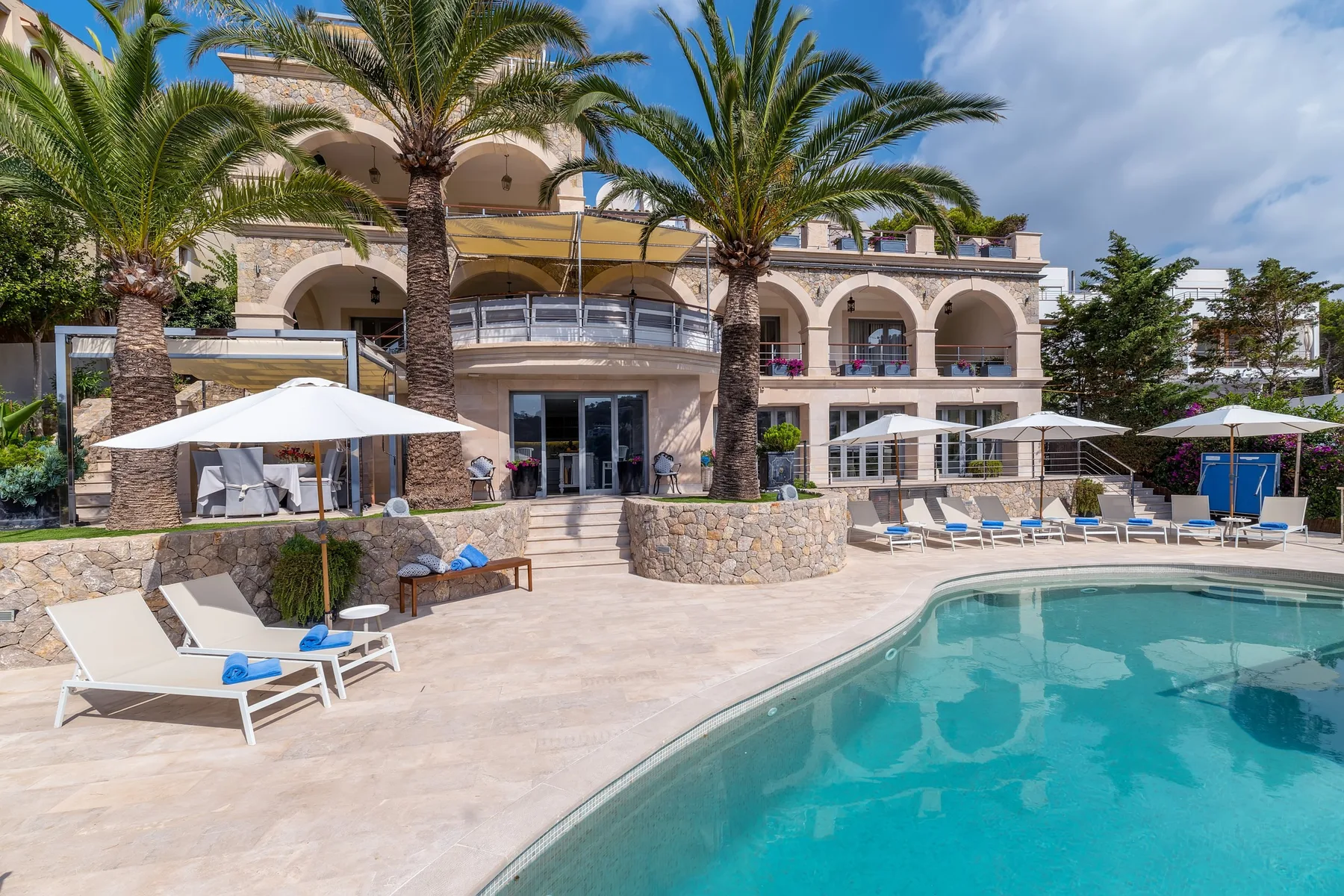 A swimming pool and sun loungers sit in front of a building with arched balconies.