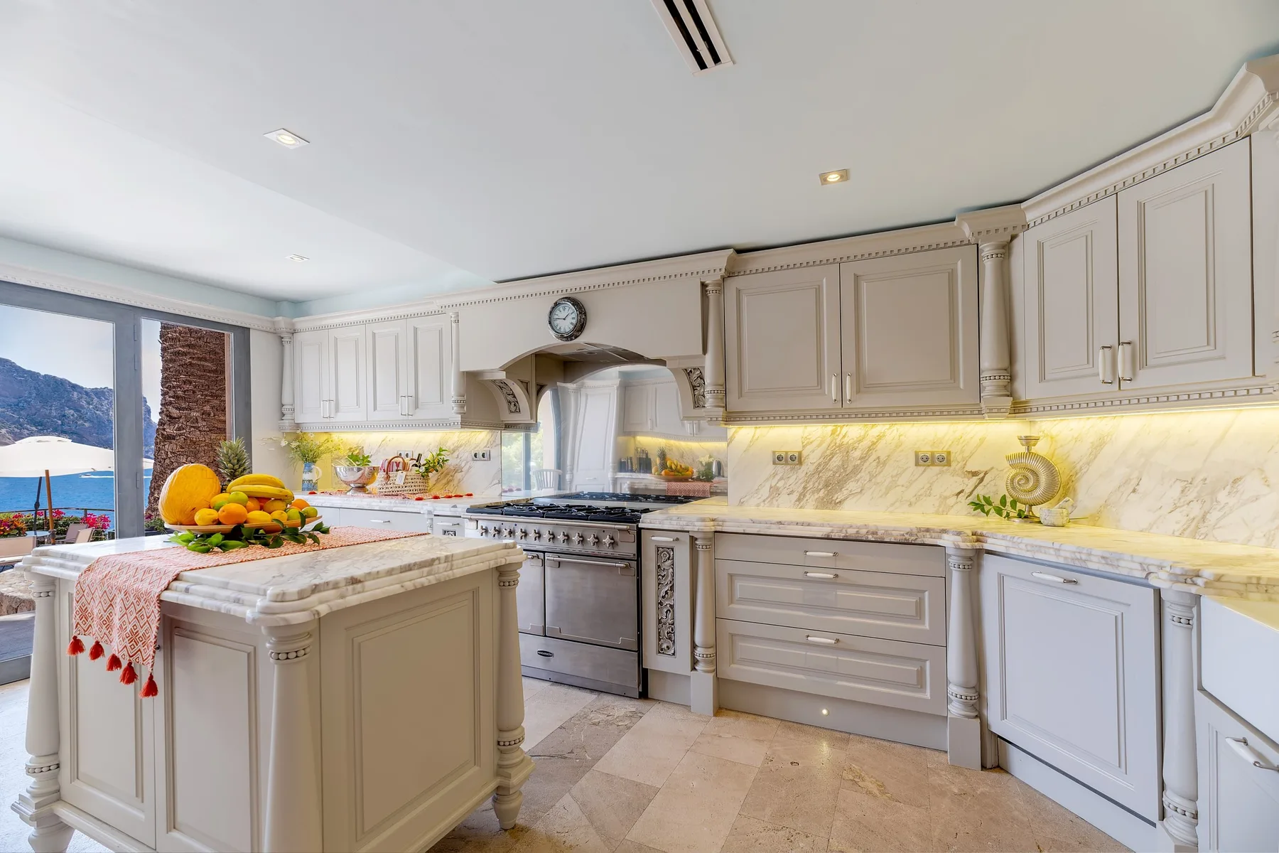 A bright kitchen with an island, pale cabinetry, and a large window overlooking the water.