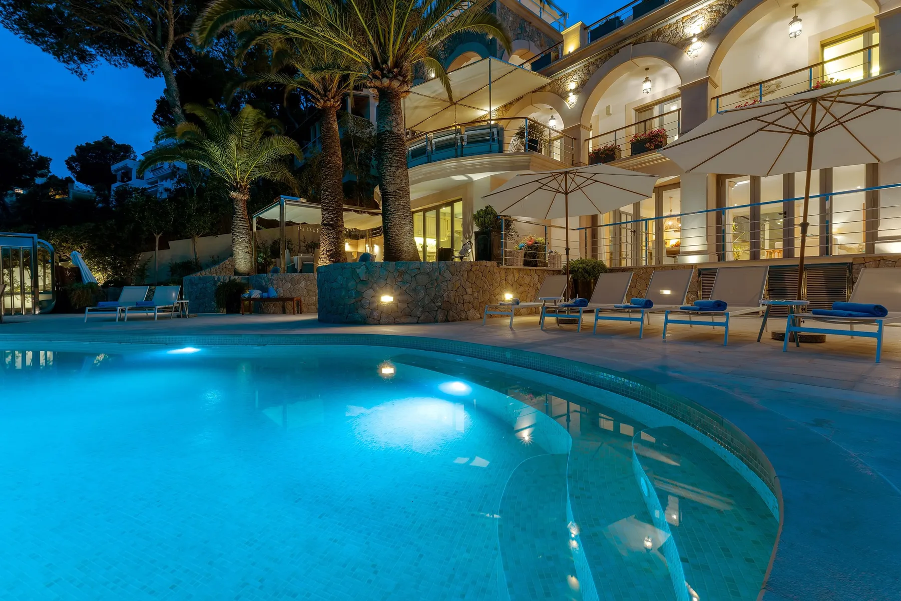 An illuminated swimming pool sits beside a multi-story building at dusk.
