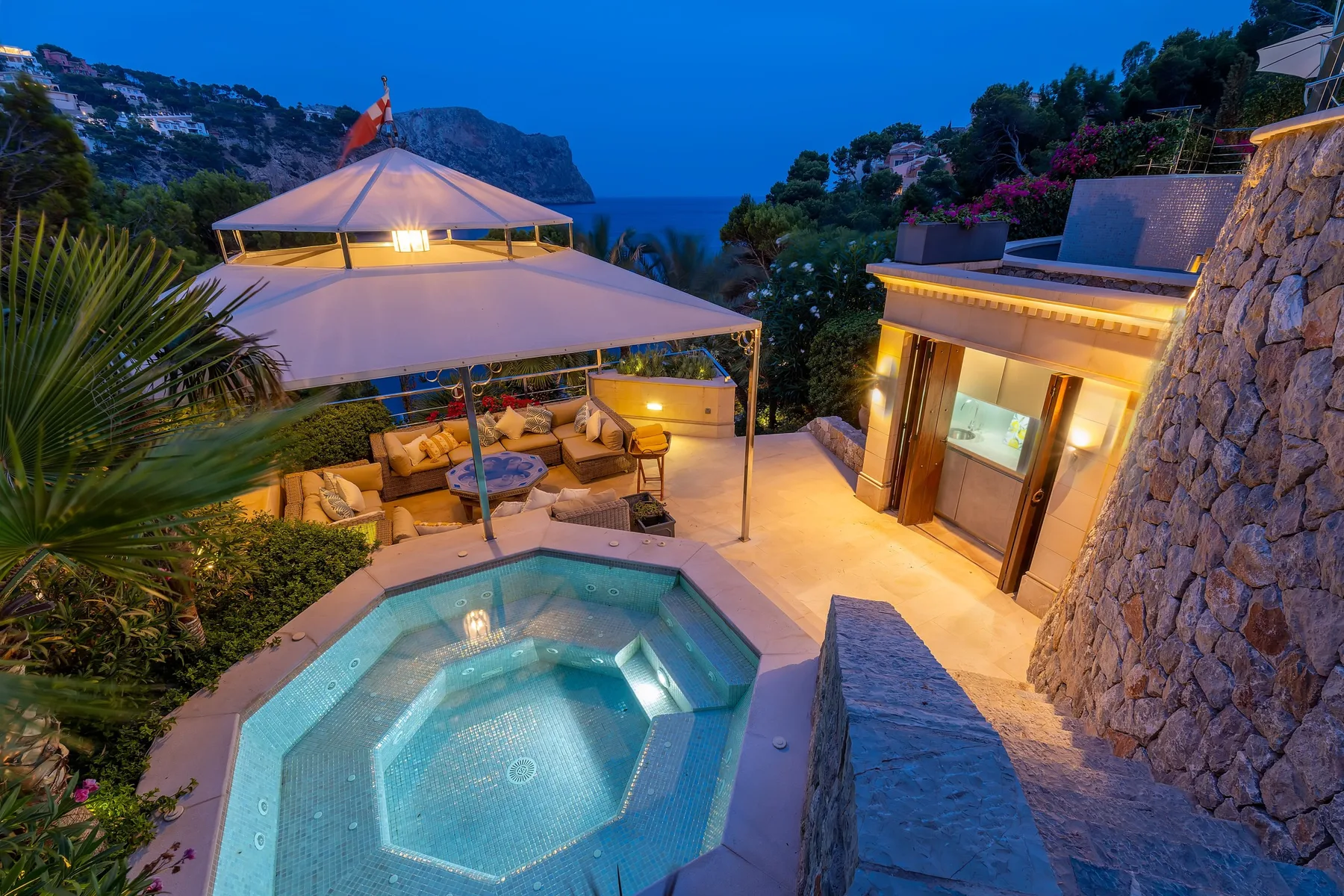 An outdoor terrace with a small illuminated pool, covered seating, and a coastal view.