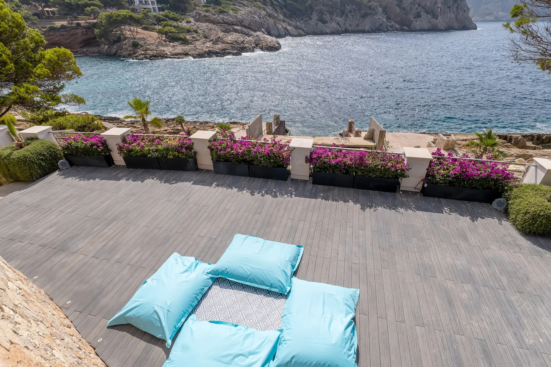 A seaside terrace with turquoise cushions and flowering planters overlooking the water.