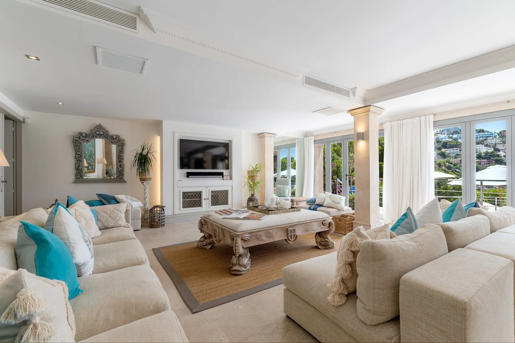 A bright living room with cushioned seating around a coffee table and views through large windows.