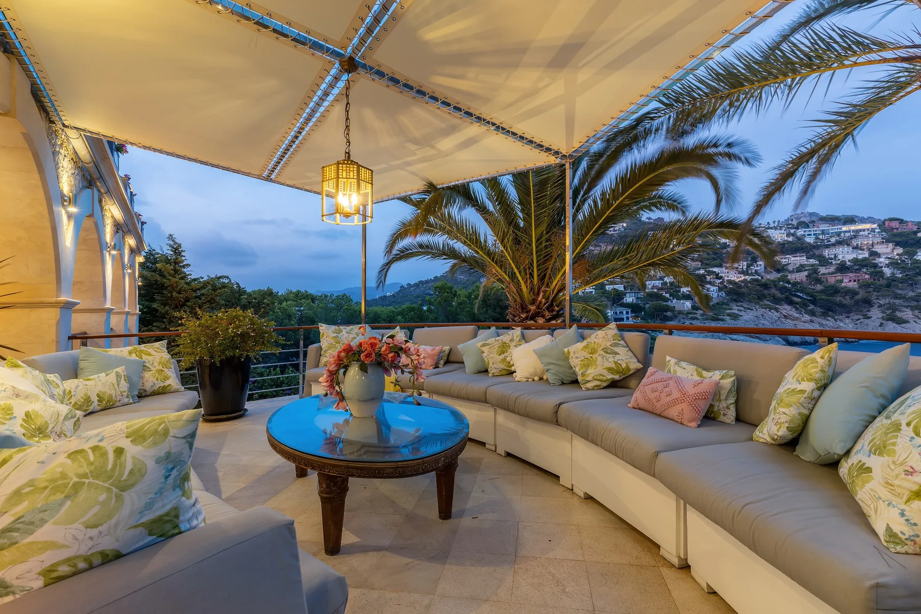 A covered outdoor patio has cushioned seating around a low table, with palm trees beyond.
