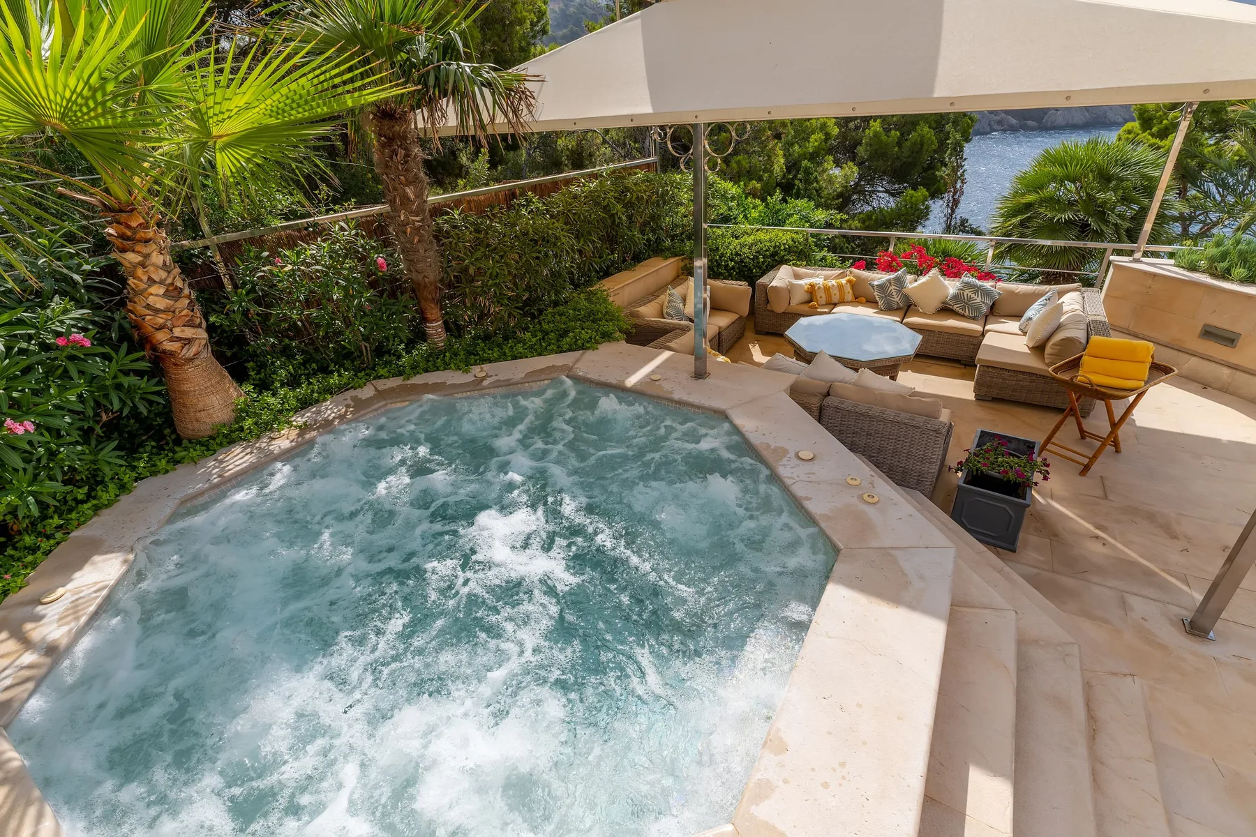 An outdoor patio features a bubbling hot tub beside cushioned seating and tropical plants.