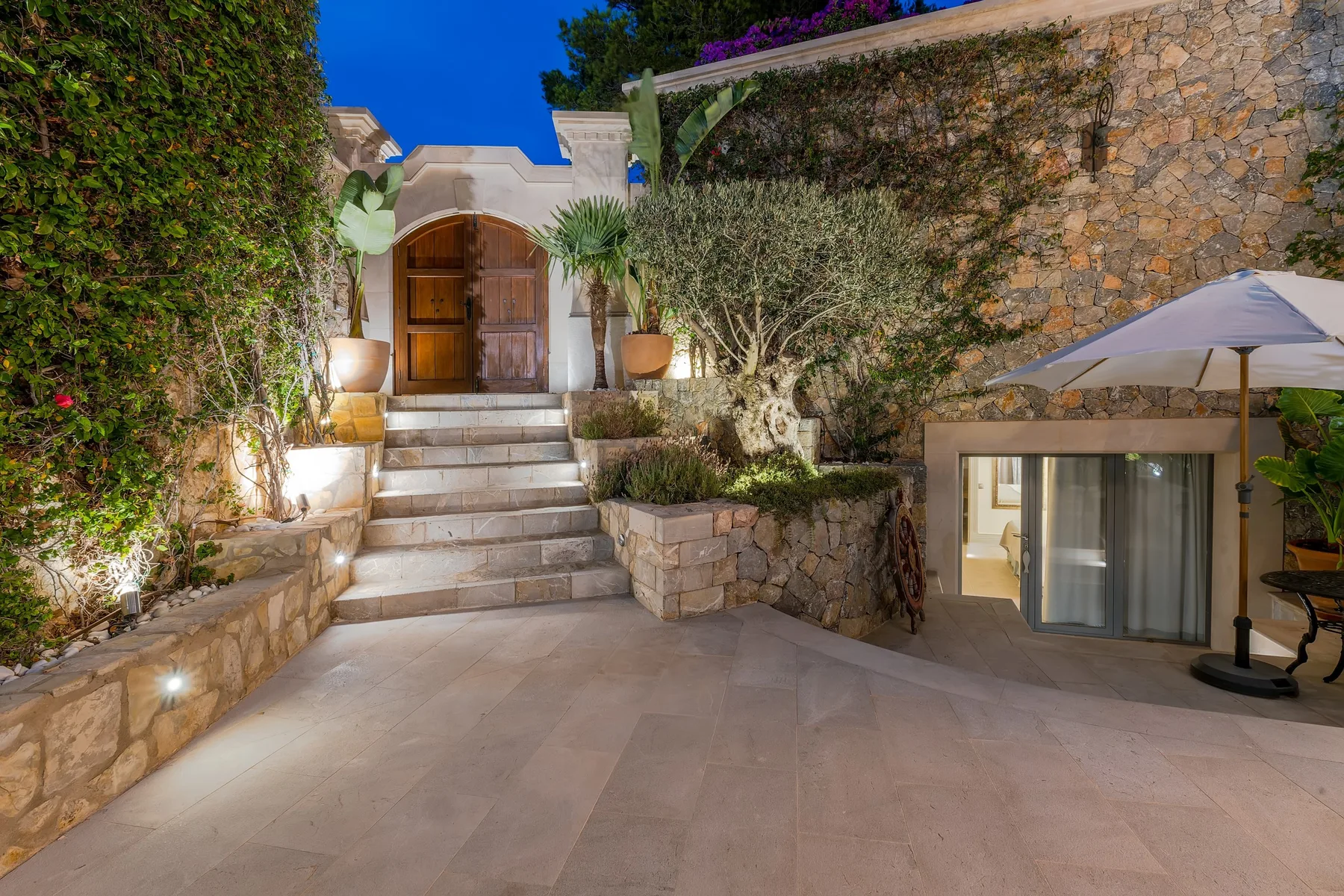 A stone courtyard with steps leading to a wooden entrance, surrounded by greenery.