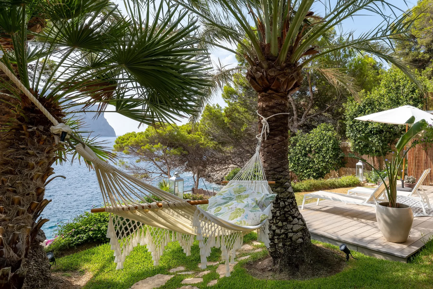 A seaside garden patio features a hammock beneath palm trees and a white umbrella.