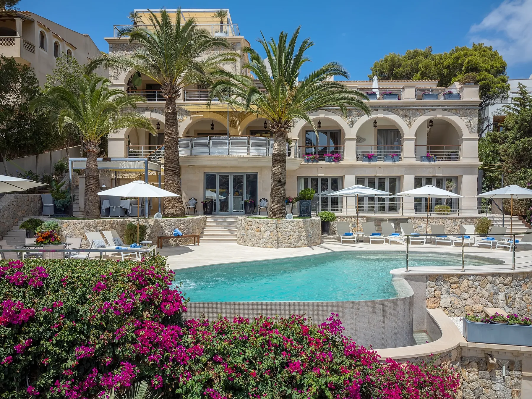 A resort building overlooks a swimming pool, with palm trees and flowering shrubs in front.