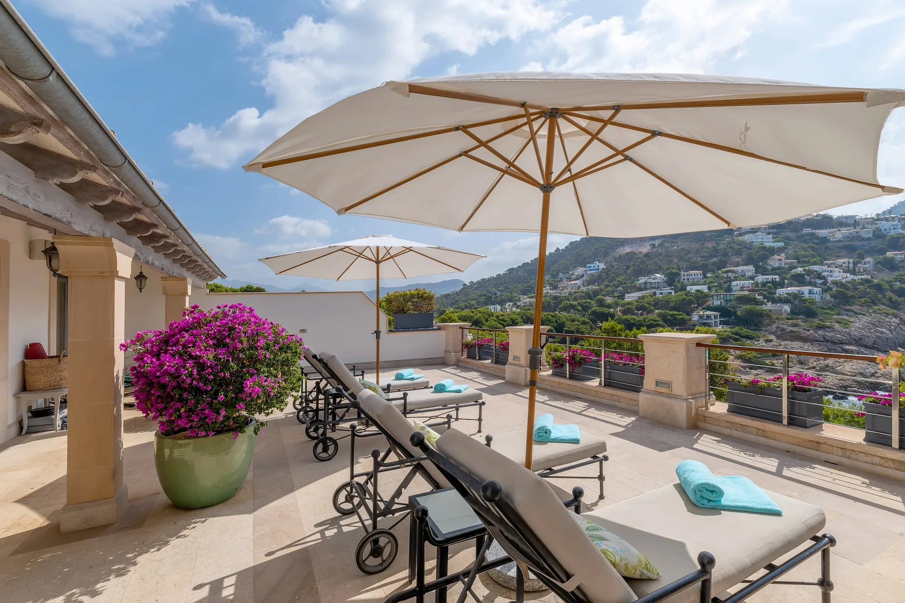 A sunny outdoor terrace with reclining seating, umbrellas, and a hillside view.
