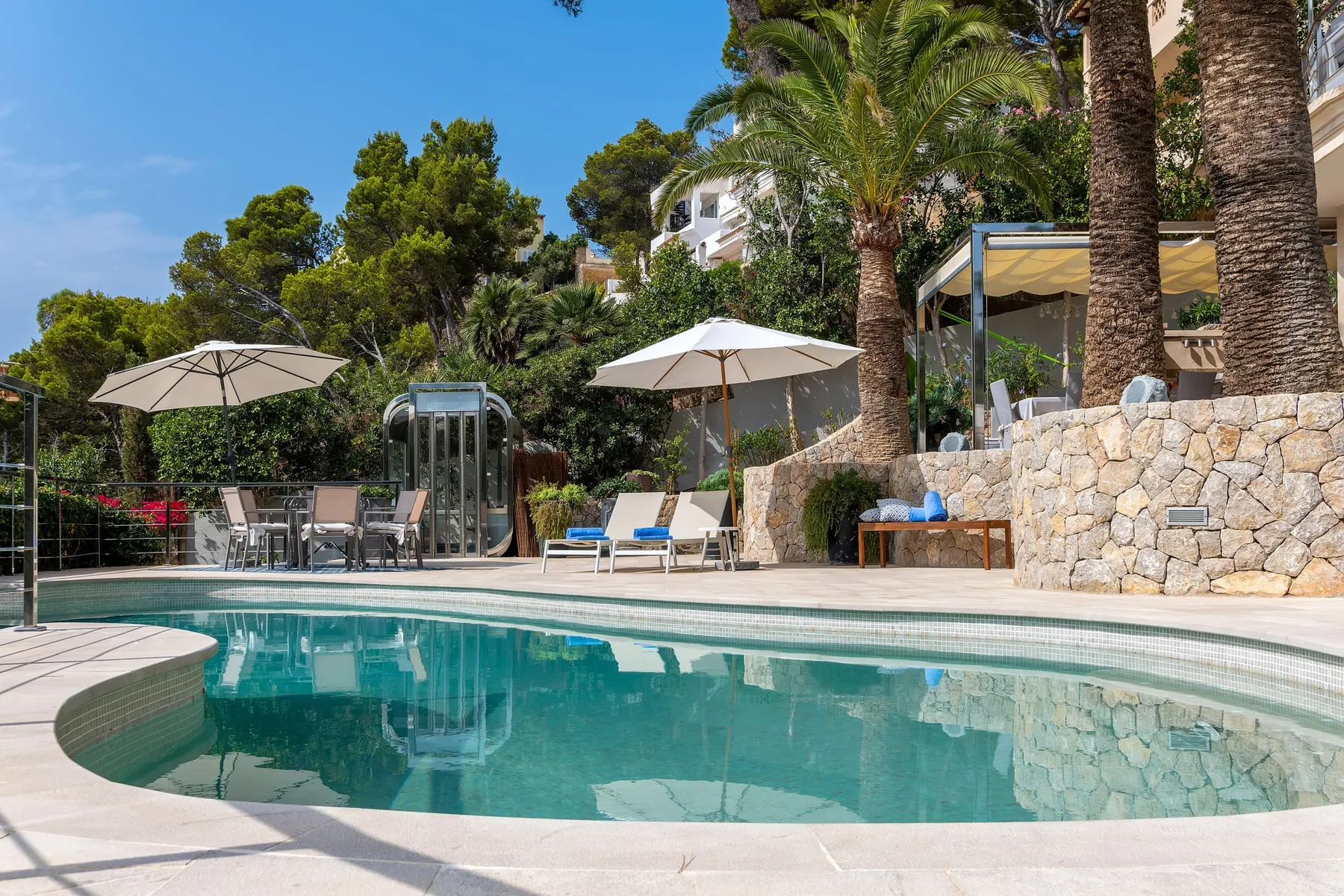 An outdoor swimming pool beside a stone wall, patio seating, and palm trees.
