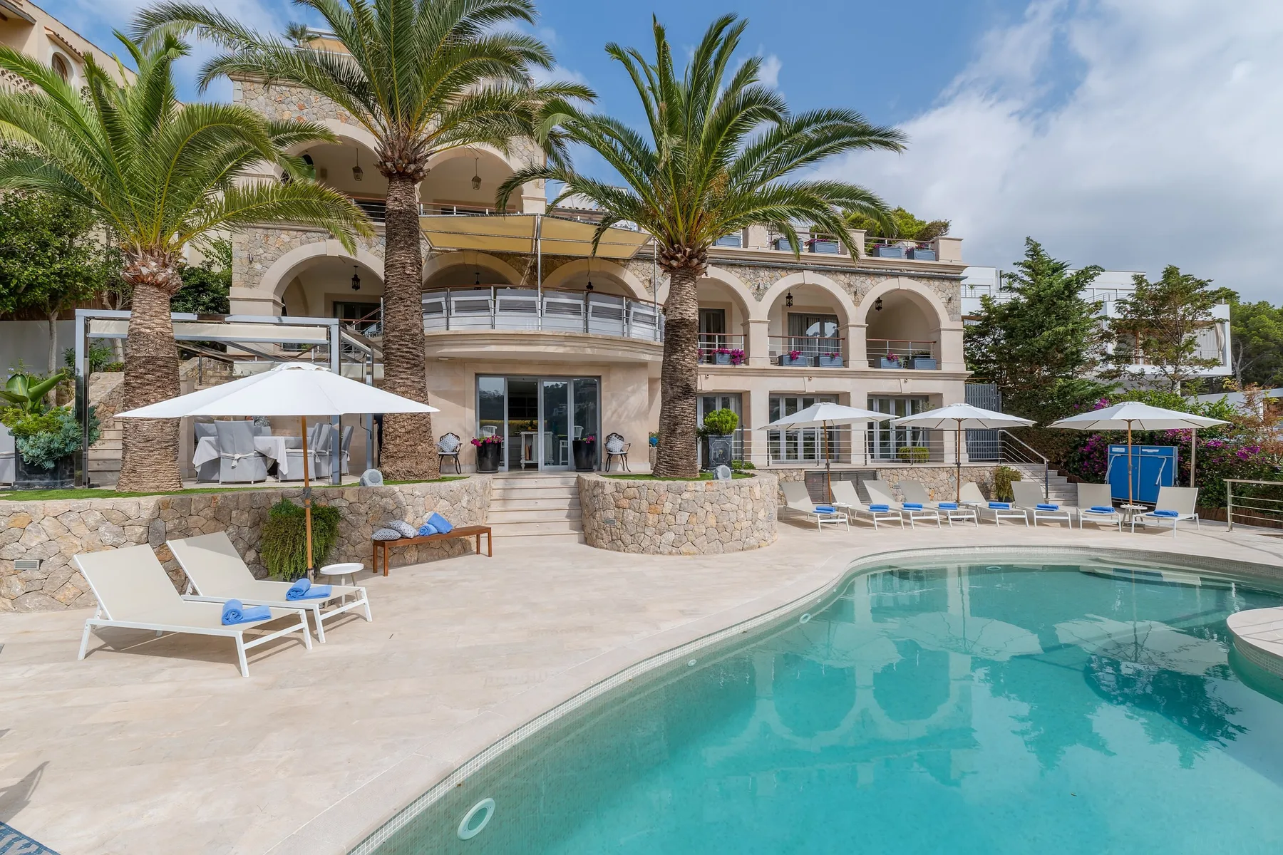 An outdoor swimming pool sits beside a palm-lined building and patio seating.