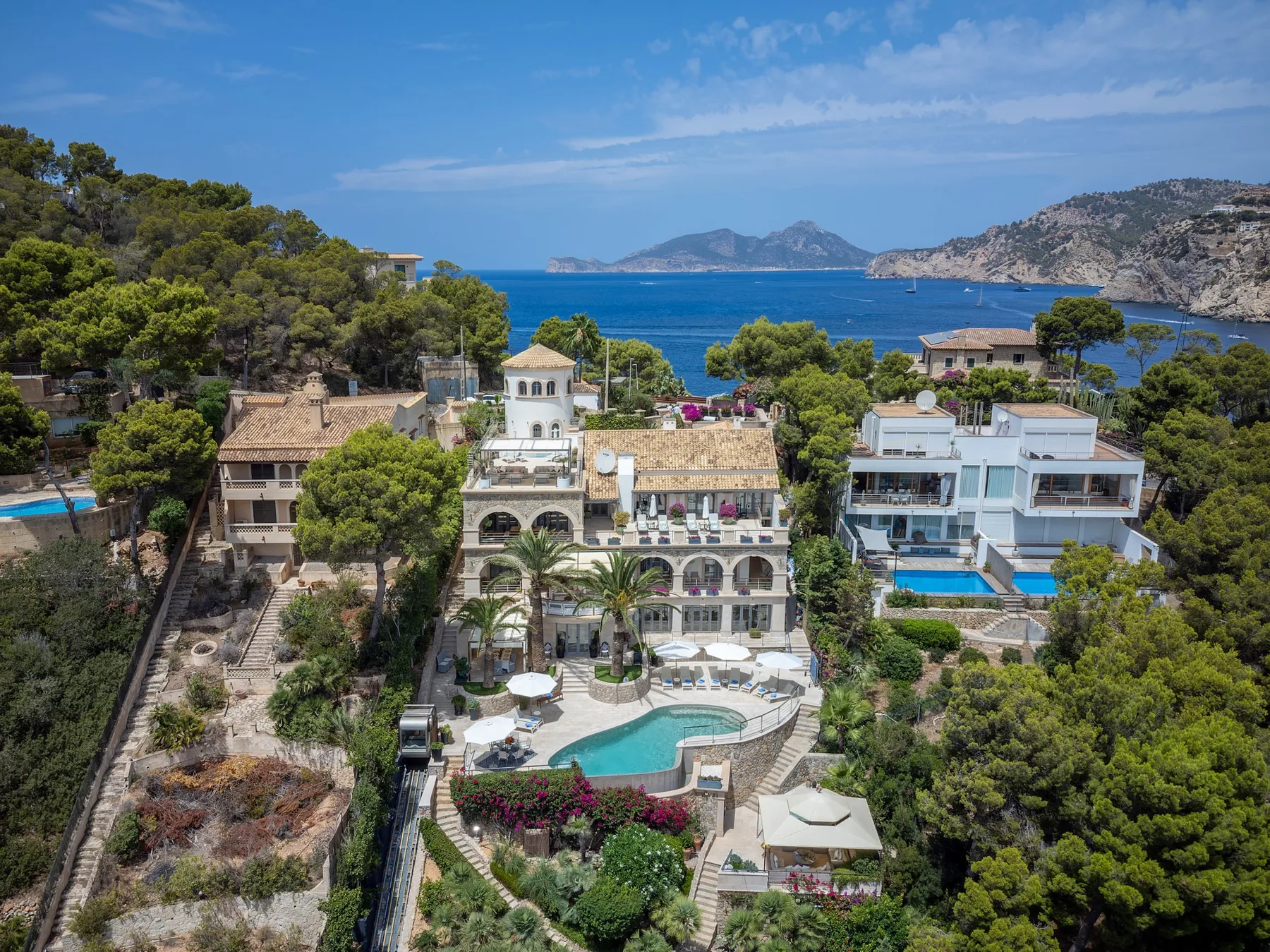 Coastal resort buildings and pools overlook a blue bay and rugged hills.