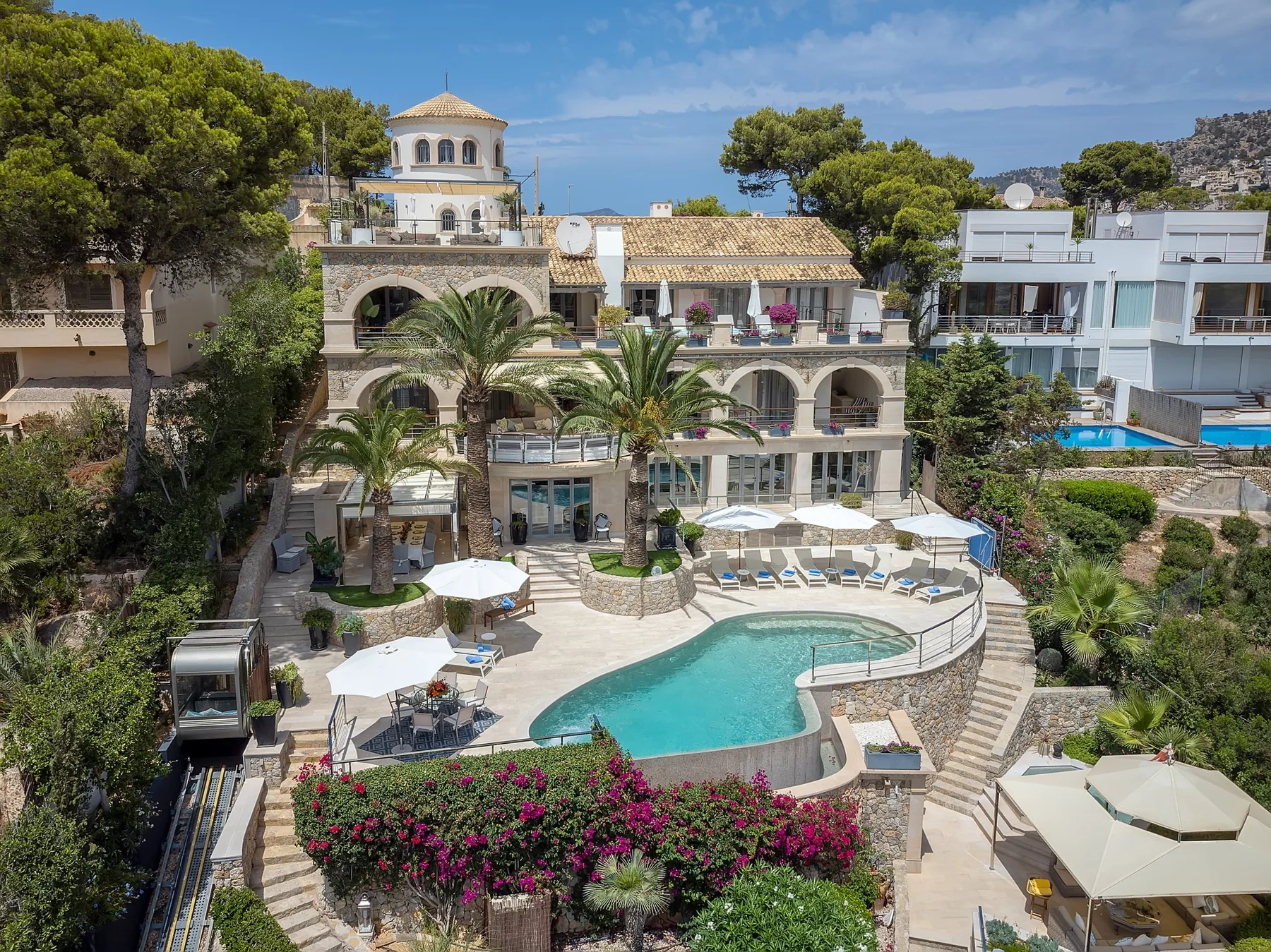A Mediterranean-style hotel courtyard with a curved pool, palm trees, and outdoor seating.
