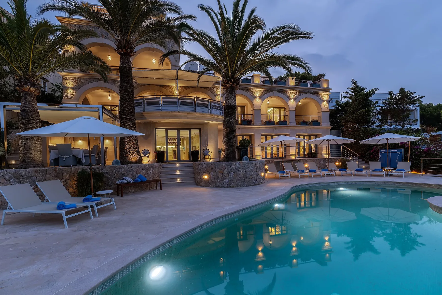 An illuminated resort building and palm trees overlook a swimming pool at dusk.