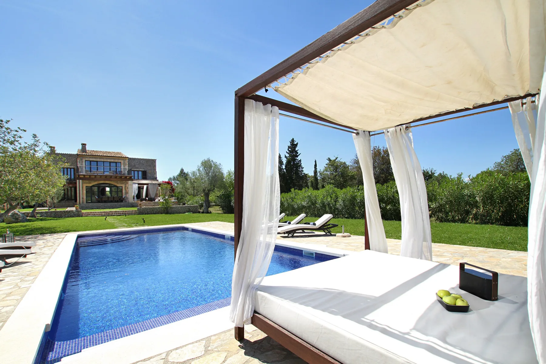 Outdoor pool beside a shaded daybed with white curtains and a lawn beyond.