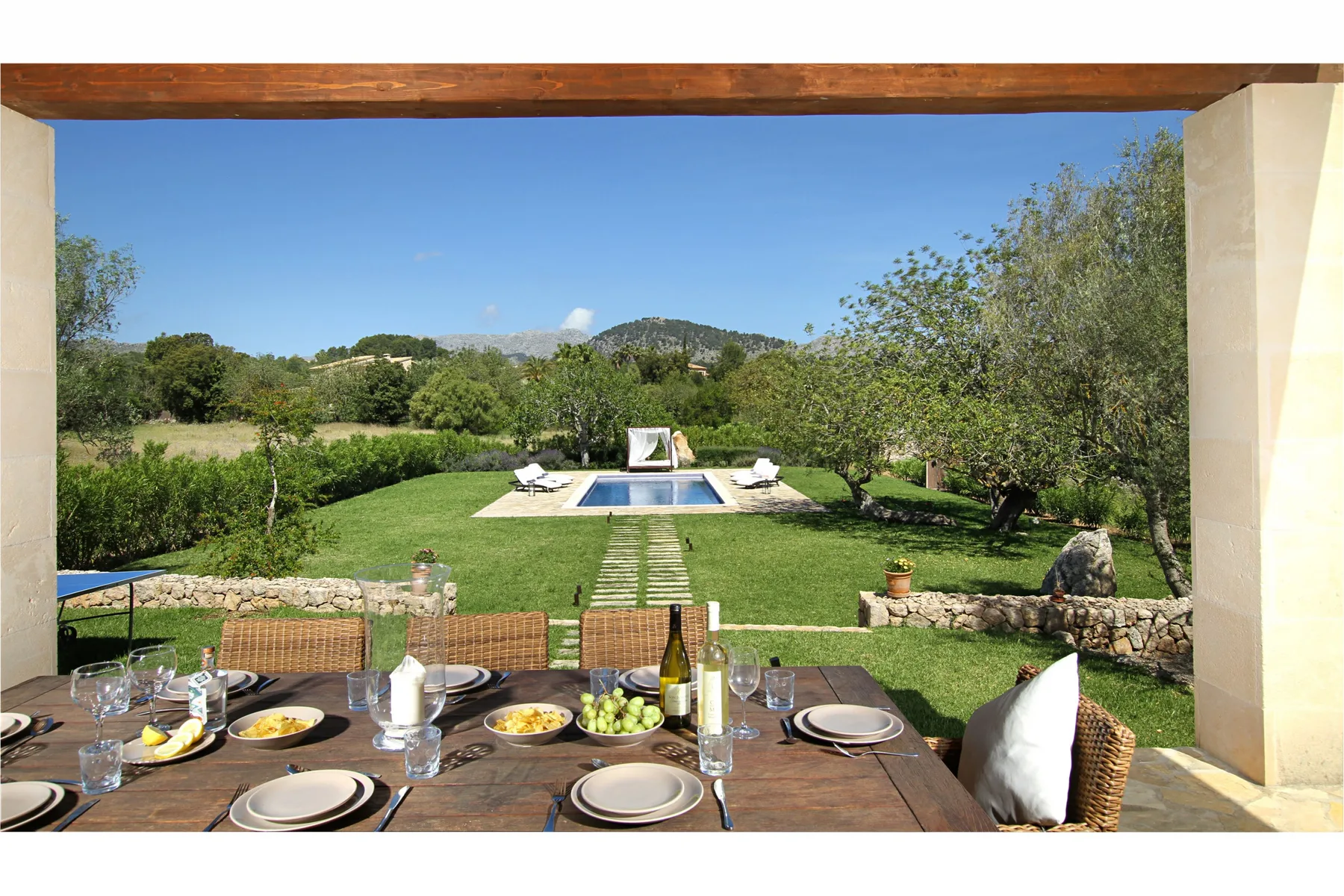 A dining table overlooks a landscaped garden with a swimming pool and distant hills.