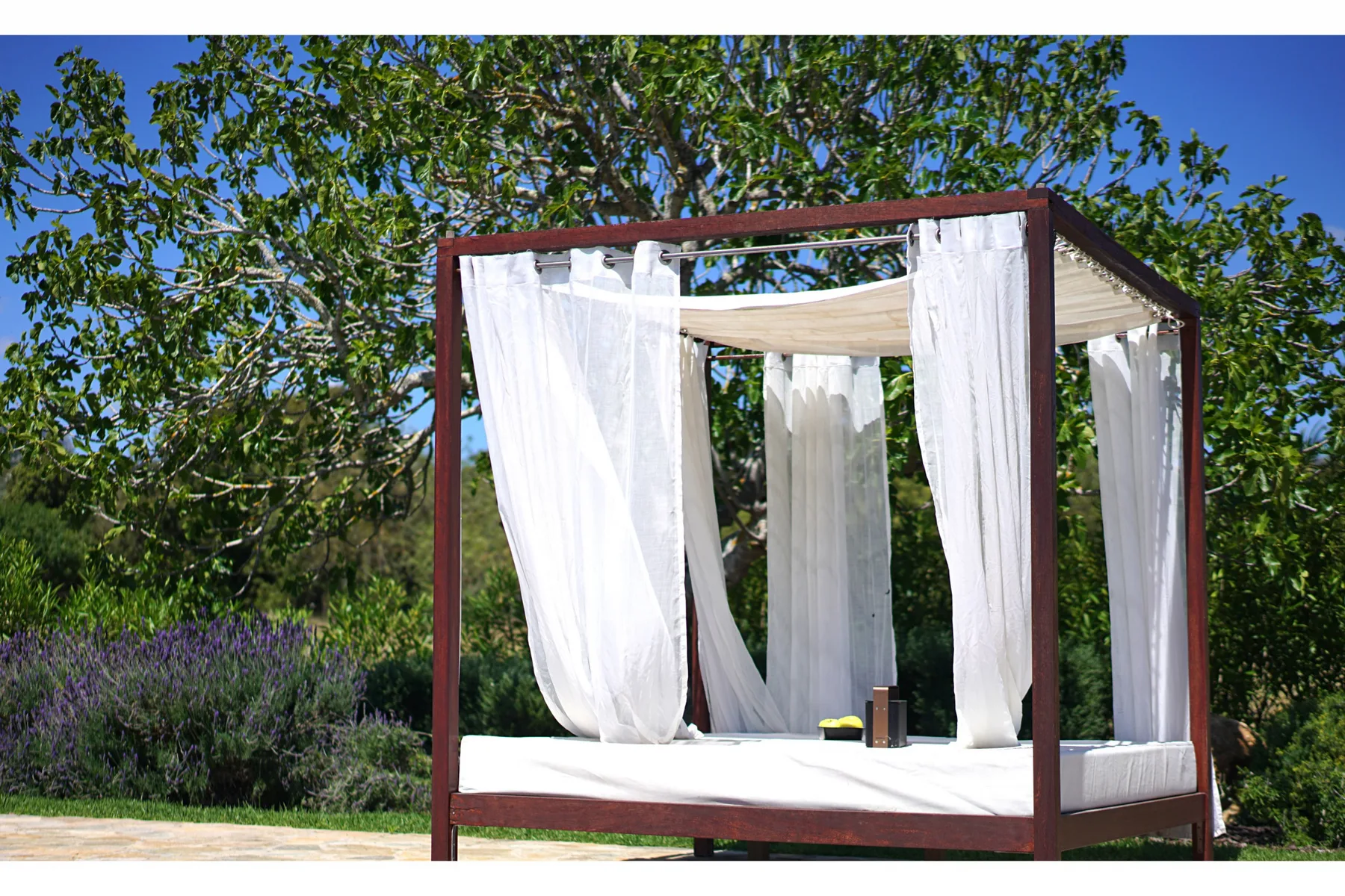 An outdoor canopy bed with white curtains sits beneath leafy trees.