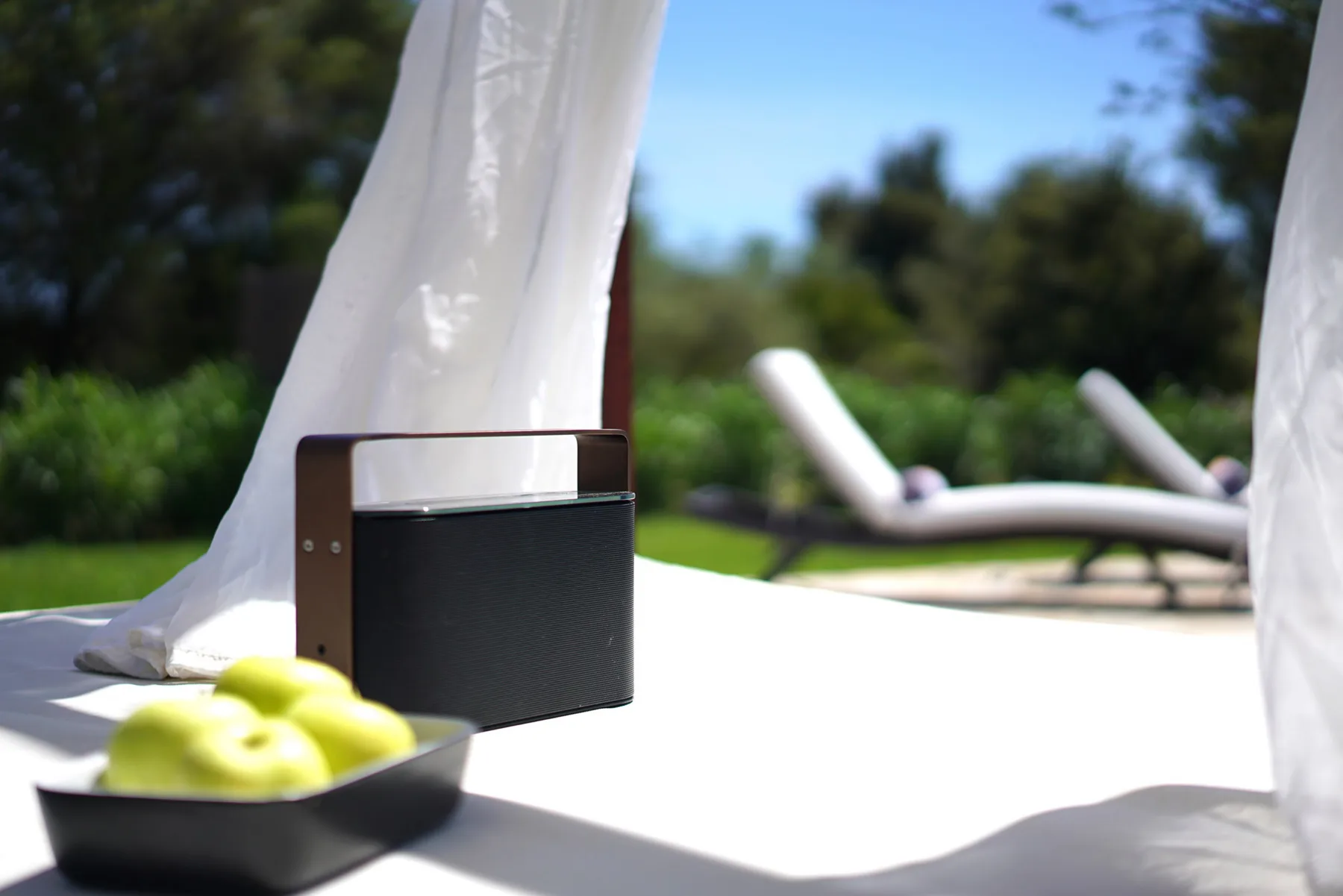 A white outdoor table holds a small speaker and a bowl of green fruit beside loungers.