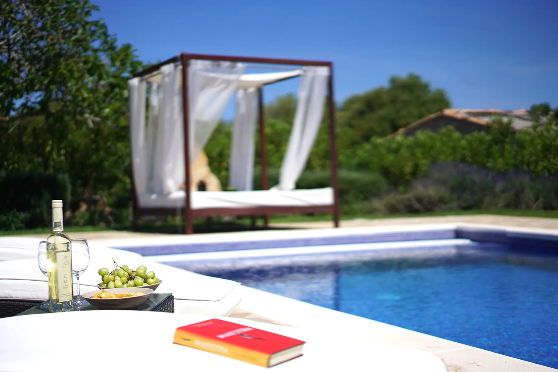 An outdoor pool beside a curtained cabana, with a bottle and fruit on a table.
