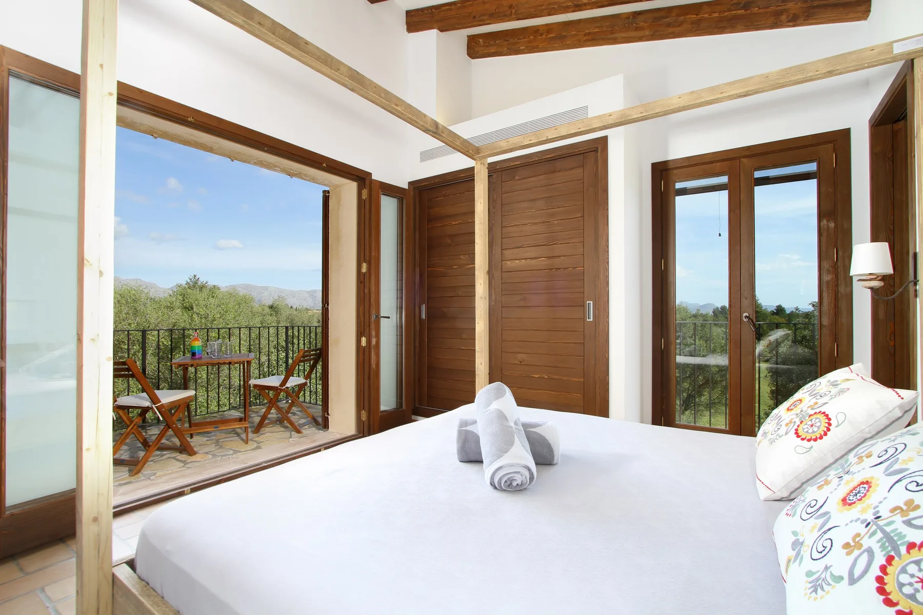 A bedroom with a bed facing wooden doors and a balcony overlooking greenery.