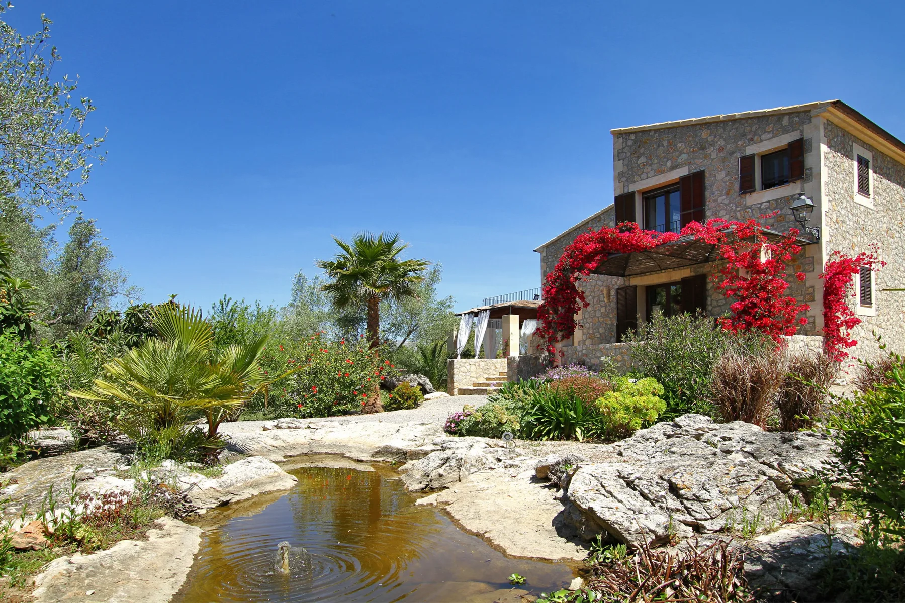 A stone house with flowering vines stands beside a rocky garden pond.