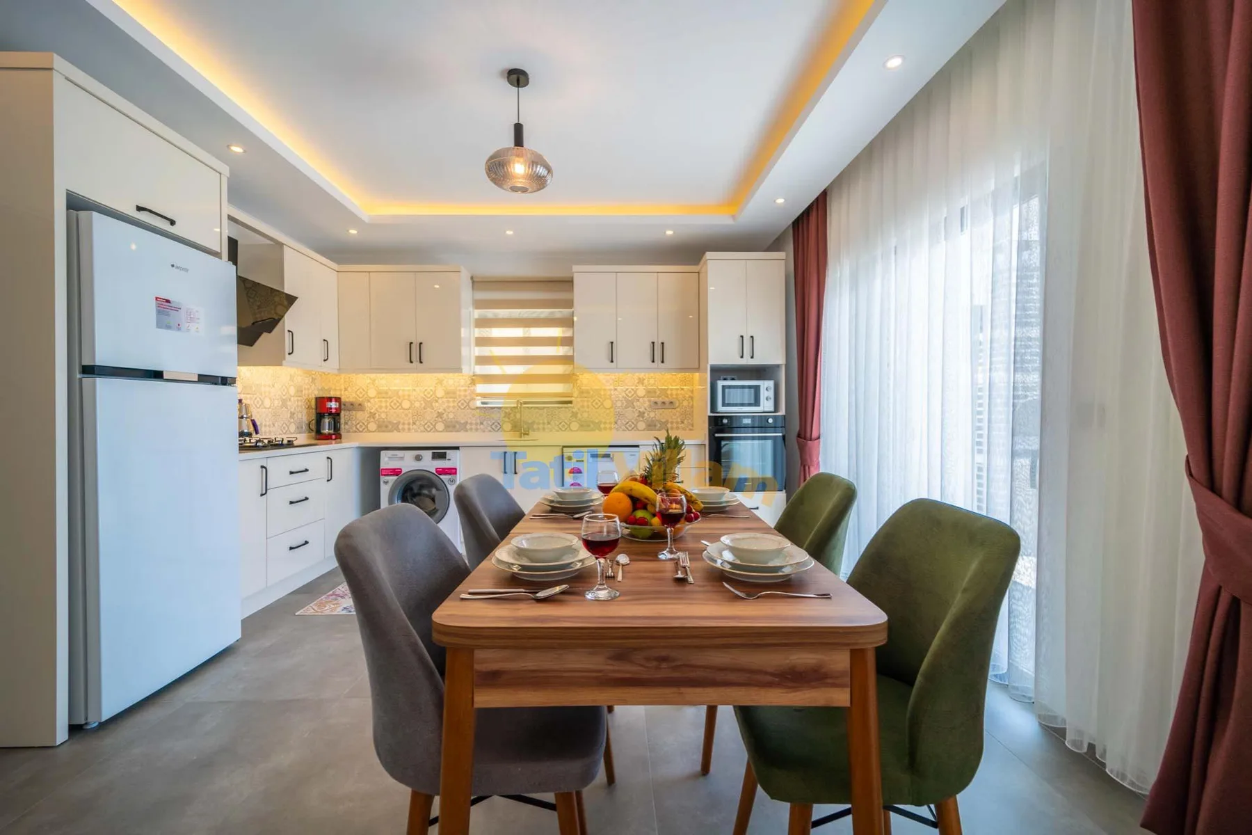 A dining area with a wooden table and upholstered seating beside a bright kitchen.