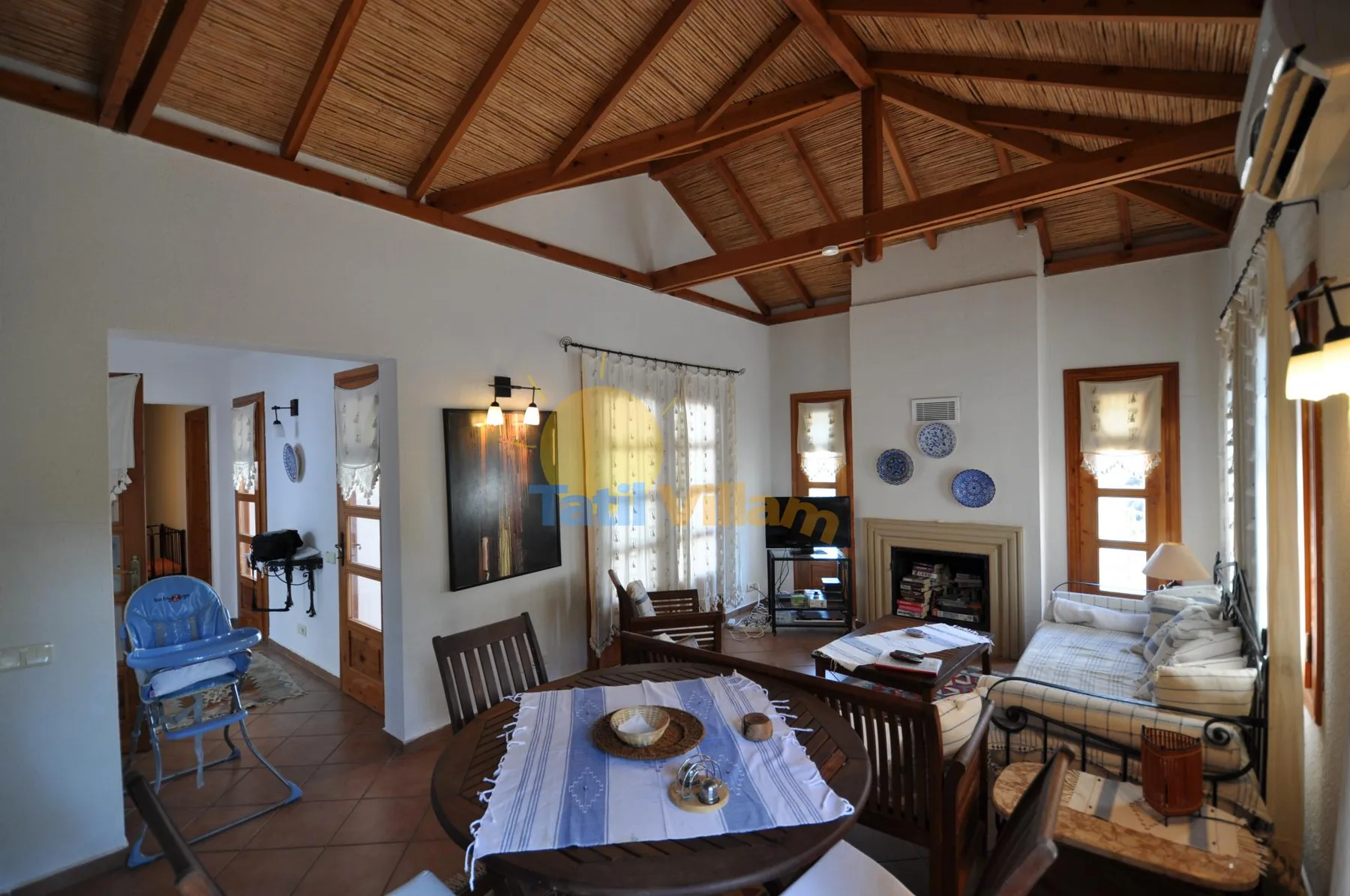 A living room with a dining table, seating, and a fireplace beneath a timber ceiling.
