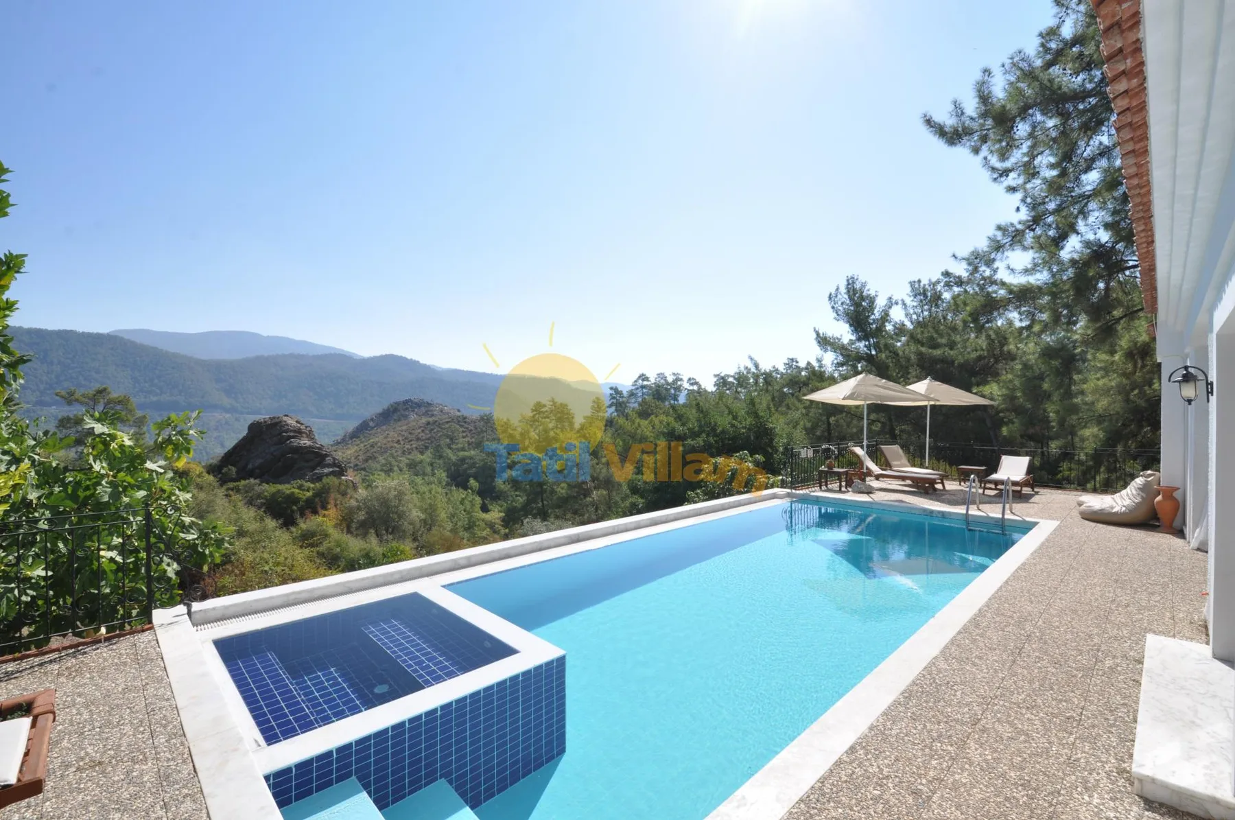 An outdoor swimming pool overlooks a wooded hillside, with a shaded seating area nearby.