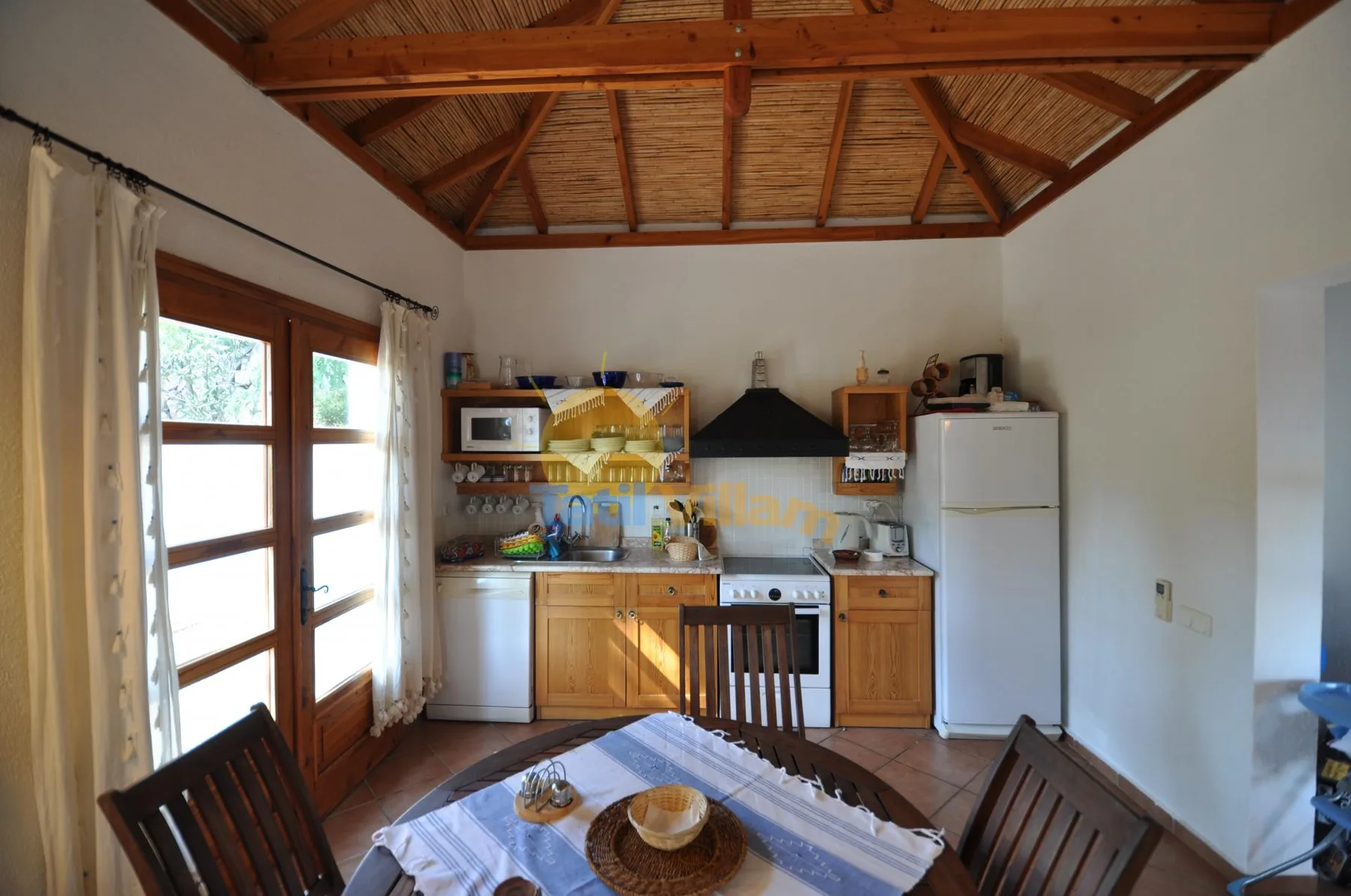 A dining area with a wooden table faces a compact kitchen beneath a timber ceiling.
