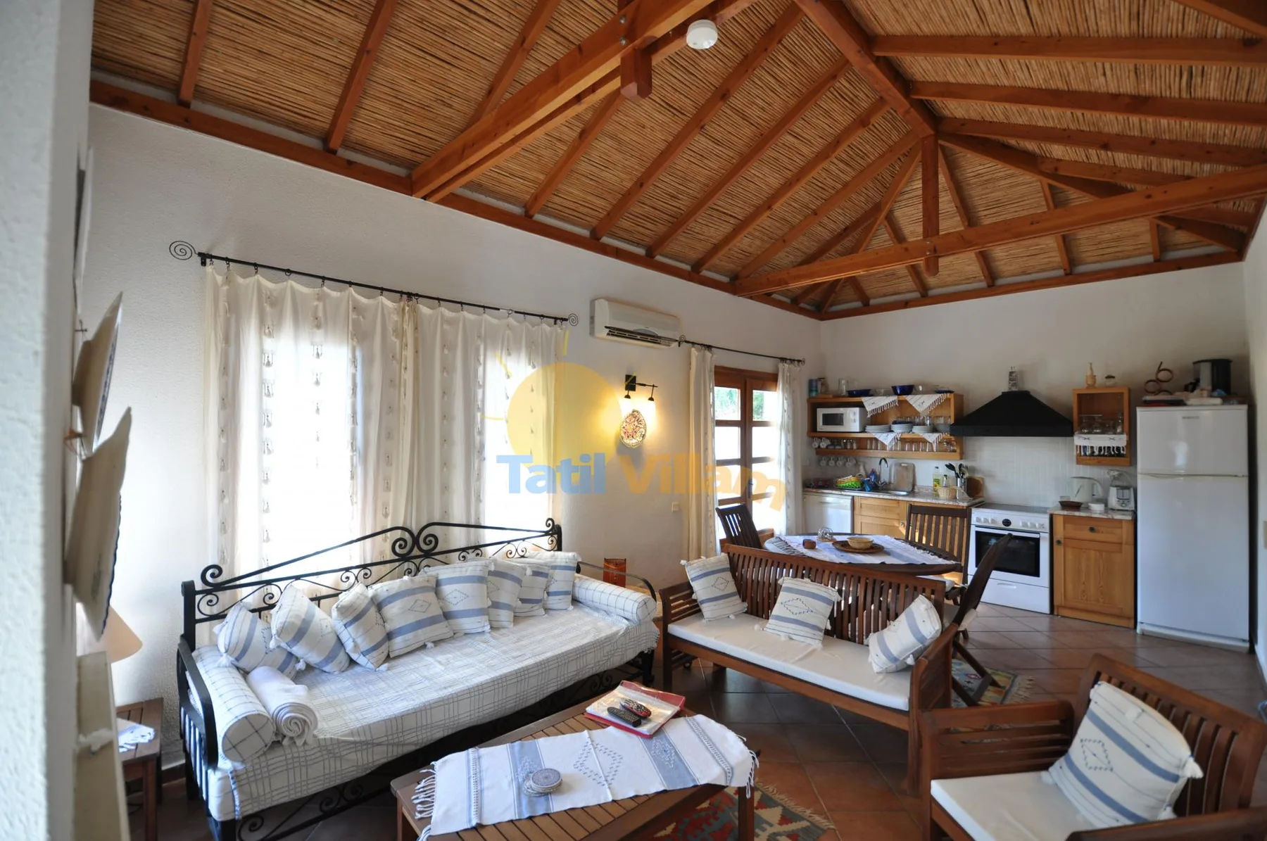 A living room with cushioned seating, a dining table, and an exposed timber ceiling.
