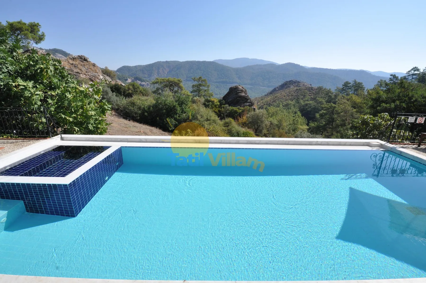 An outdoor swimming pool sits beneath a clear sky with wooded hills in the distance.