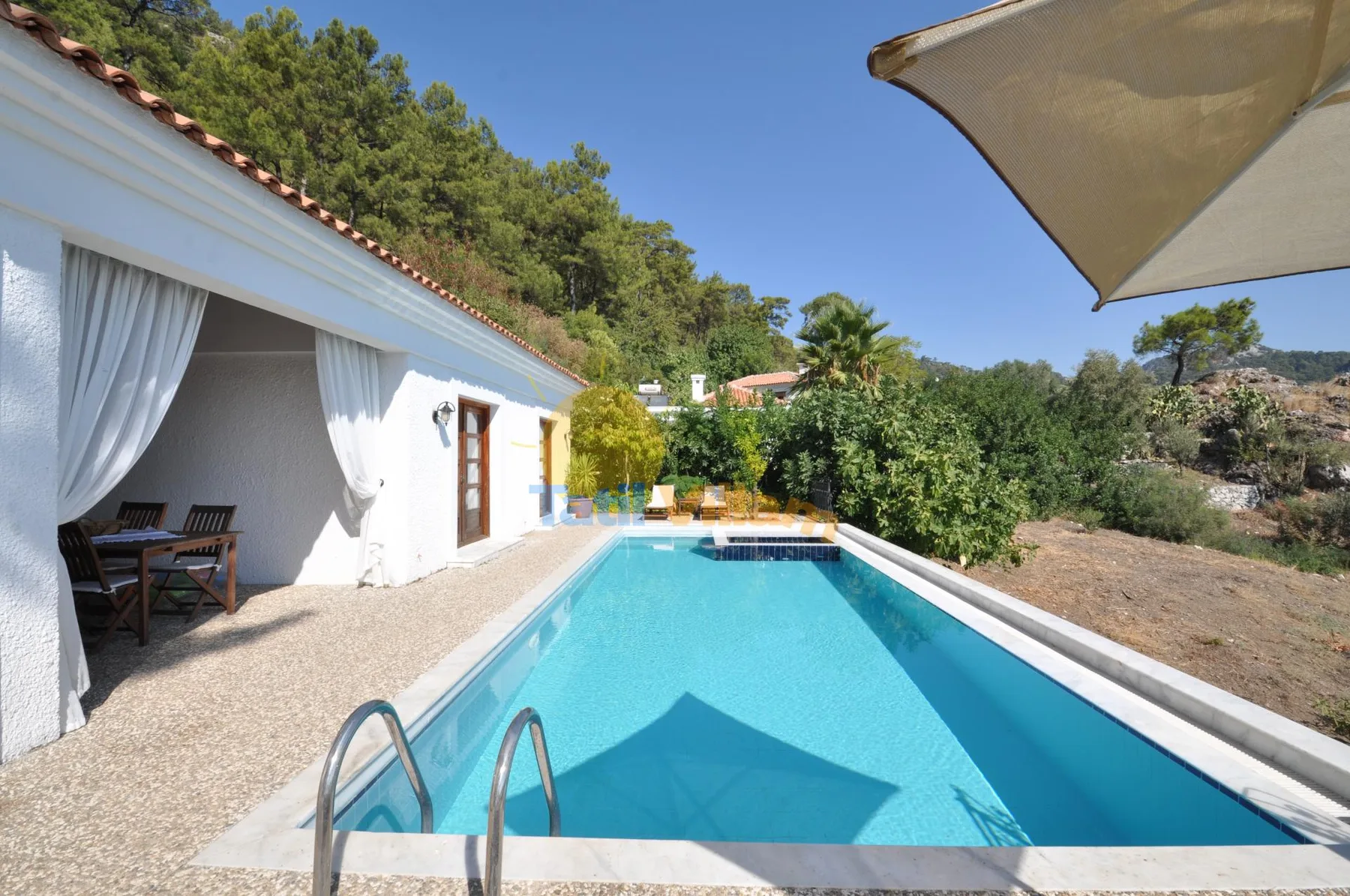 An outdoor swimming pool beside a white building, with greenery in the background.