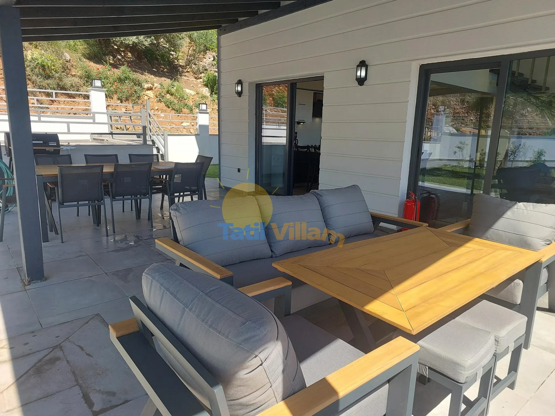 Un patio exterior cubierto tiene asientos acolchados y una mesa de comedor de madera.