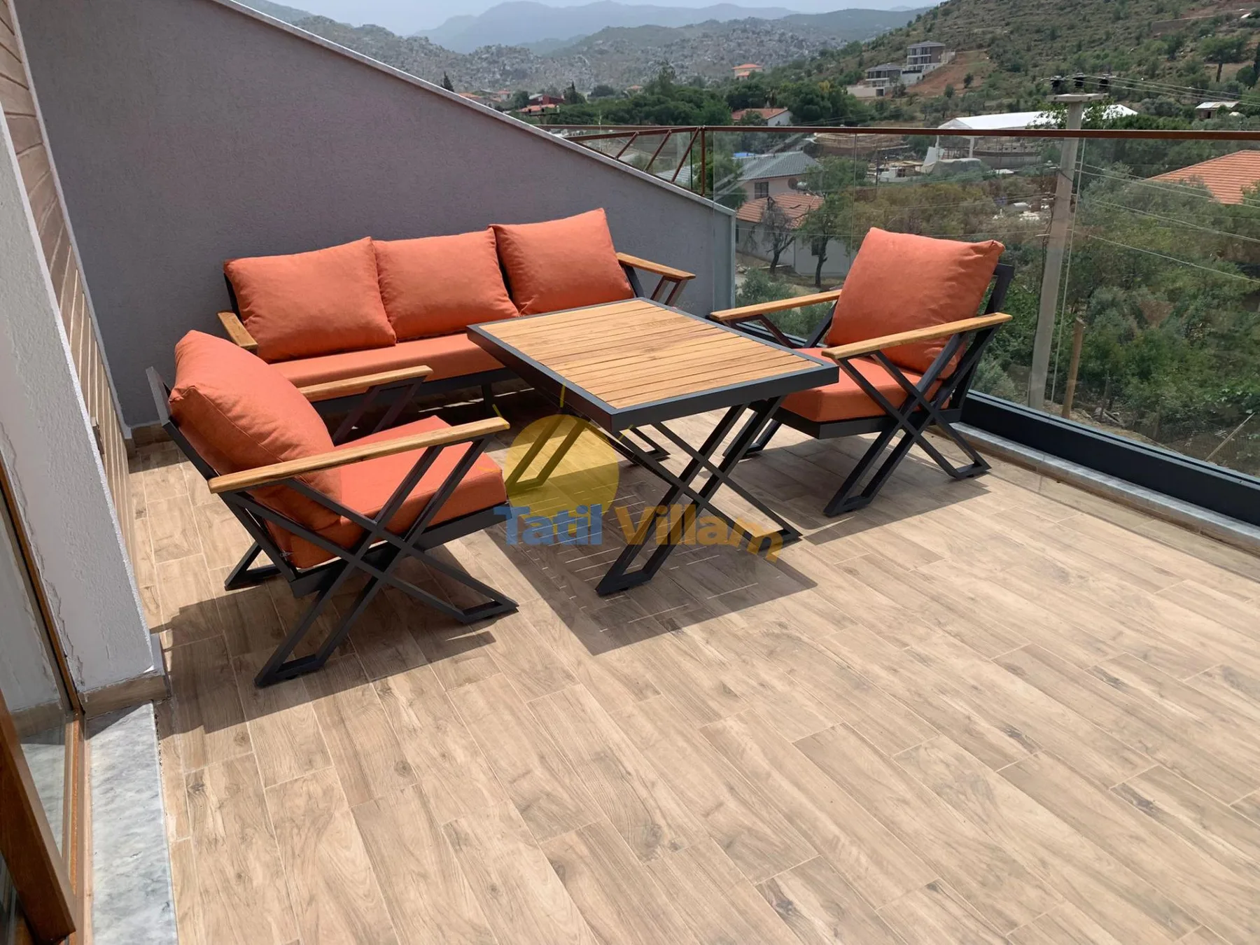 An outdoor terrace has orange-cushioned seating around a wooden table, with a hillside view.